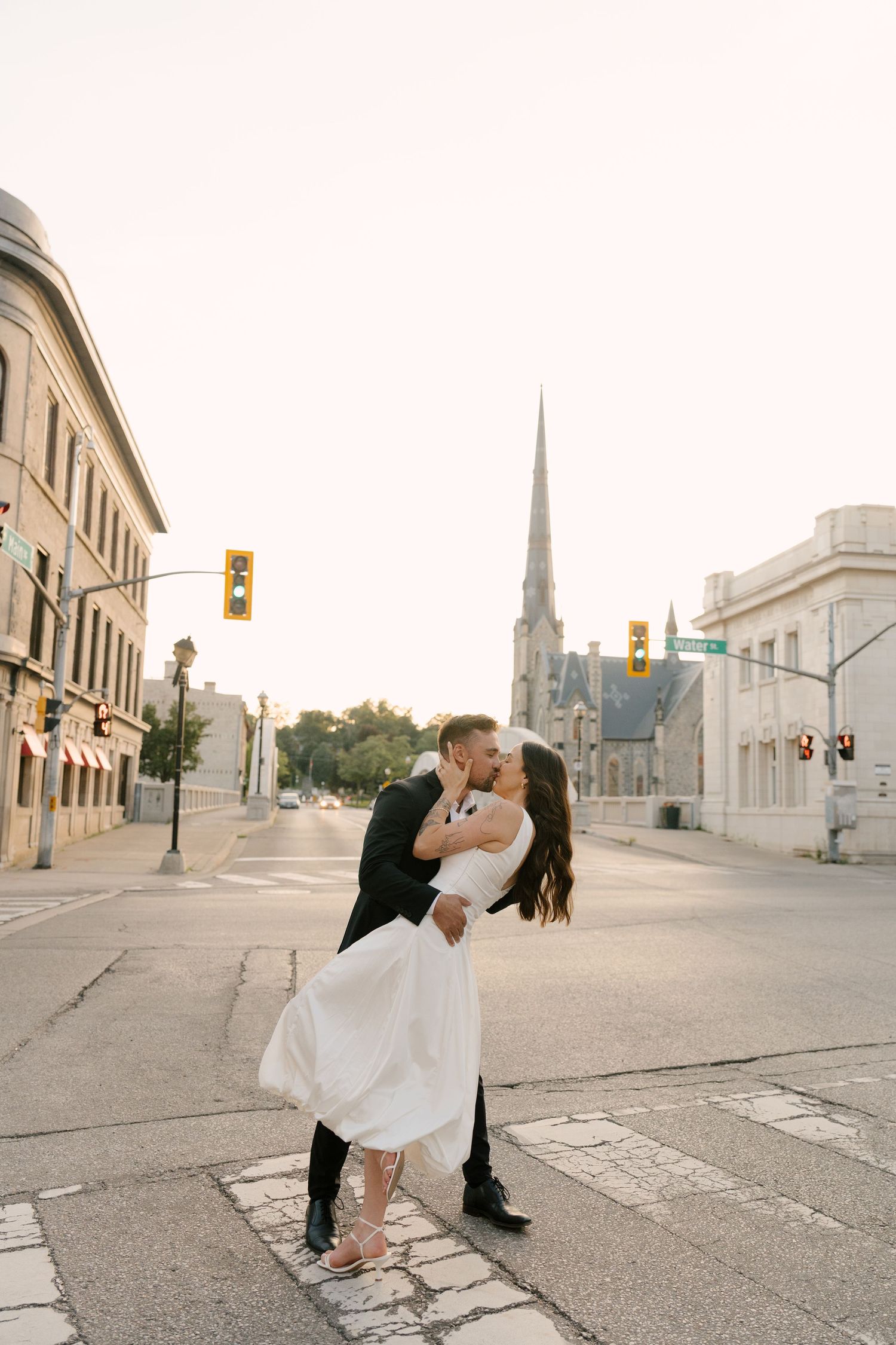 Groom dipping bride for a kiss at a city intersection with church steeple in background.
