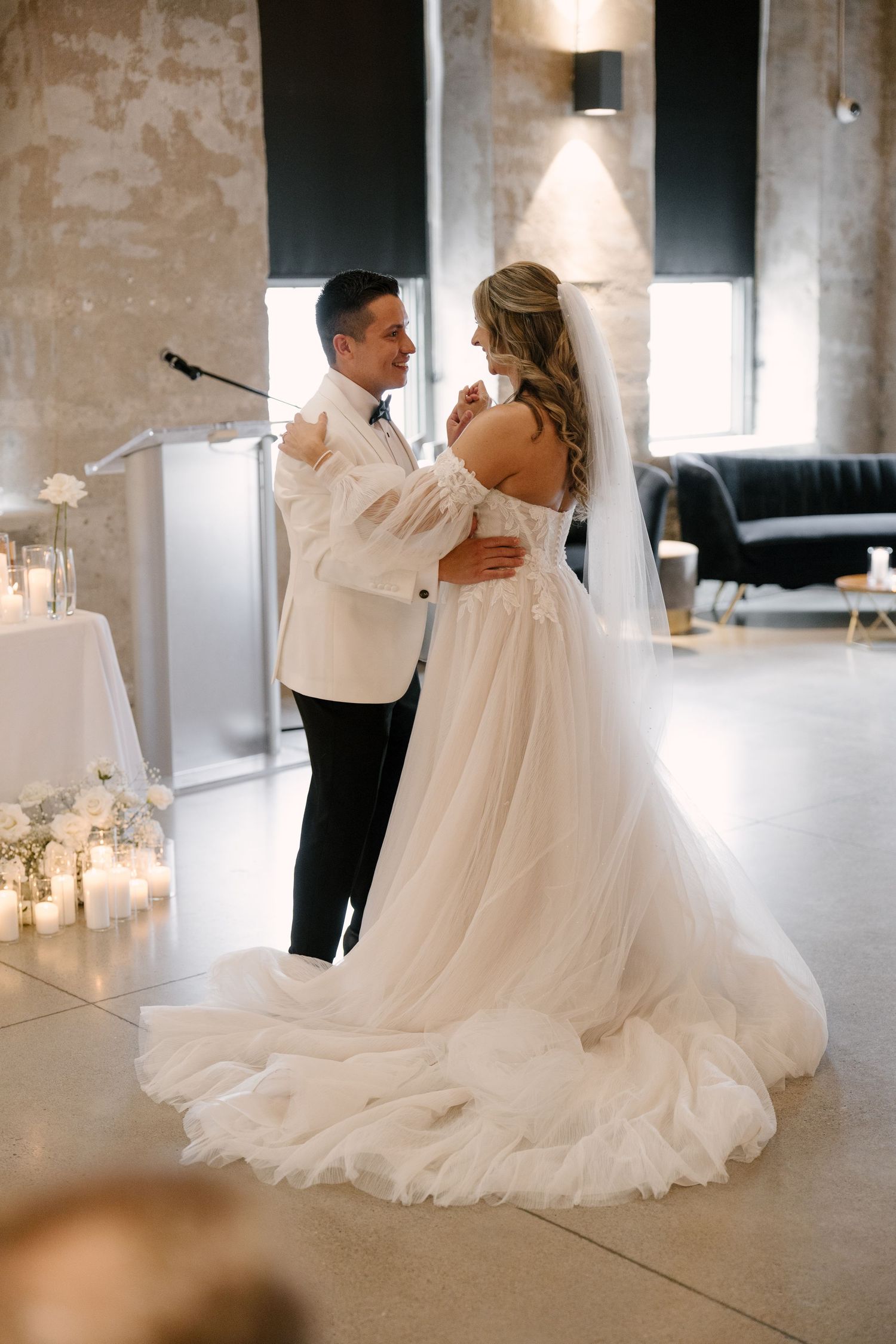 Bride in off-shoulder tulle gown and groom in white tuxedo share first dance in candlelit industrial Tapestry Hall venue.