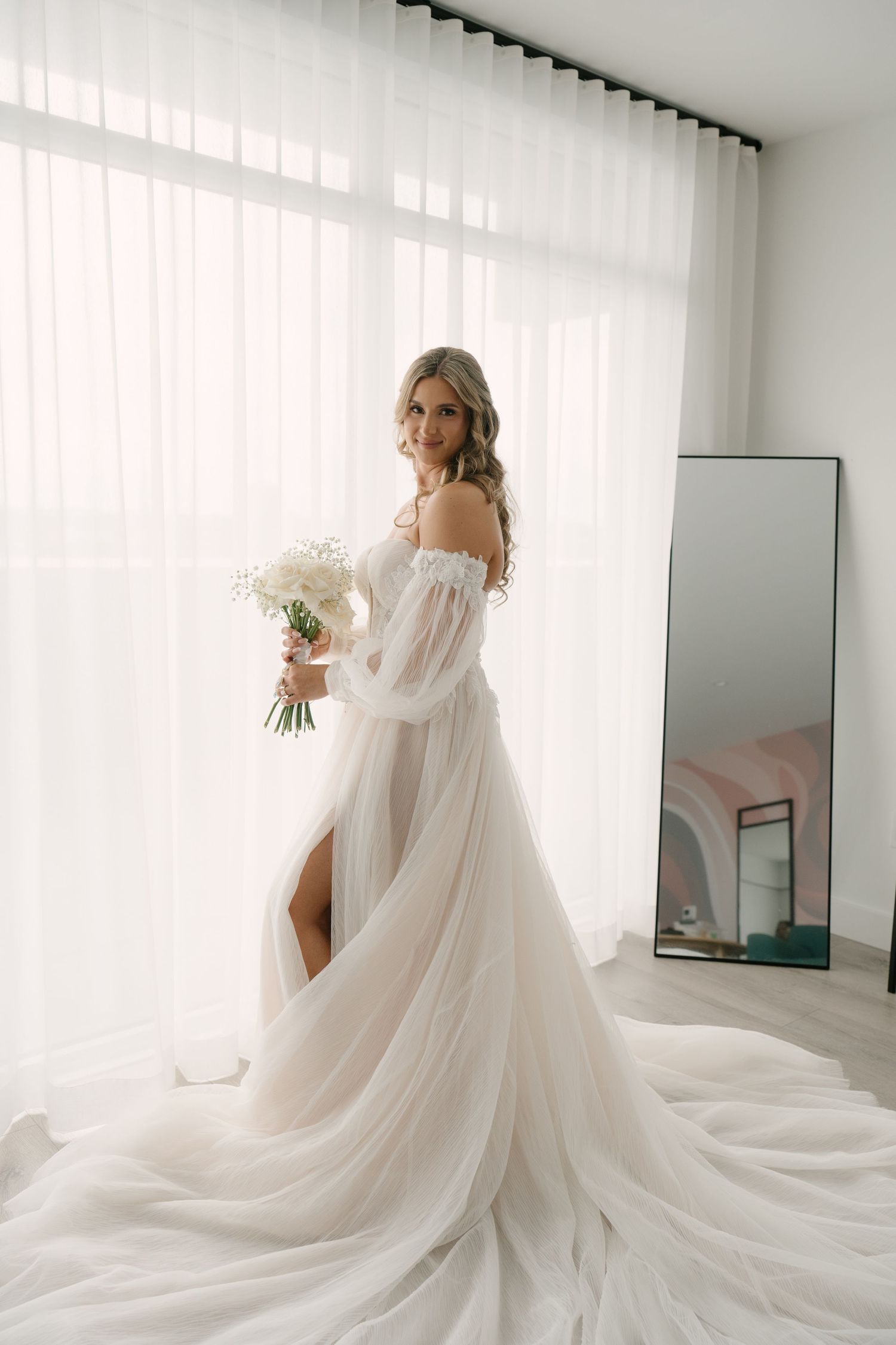 Elegant bride in off-shoulder white tulle gown holds bouquet while posing near sheer curtained window.