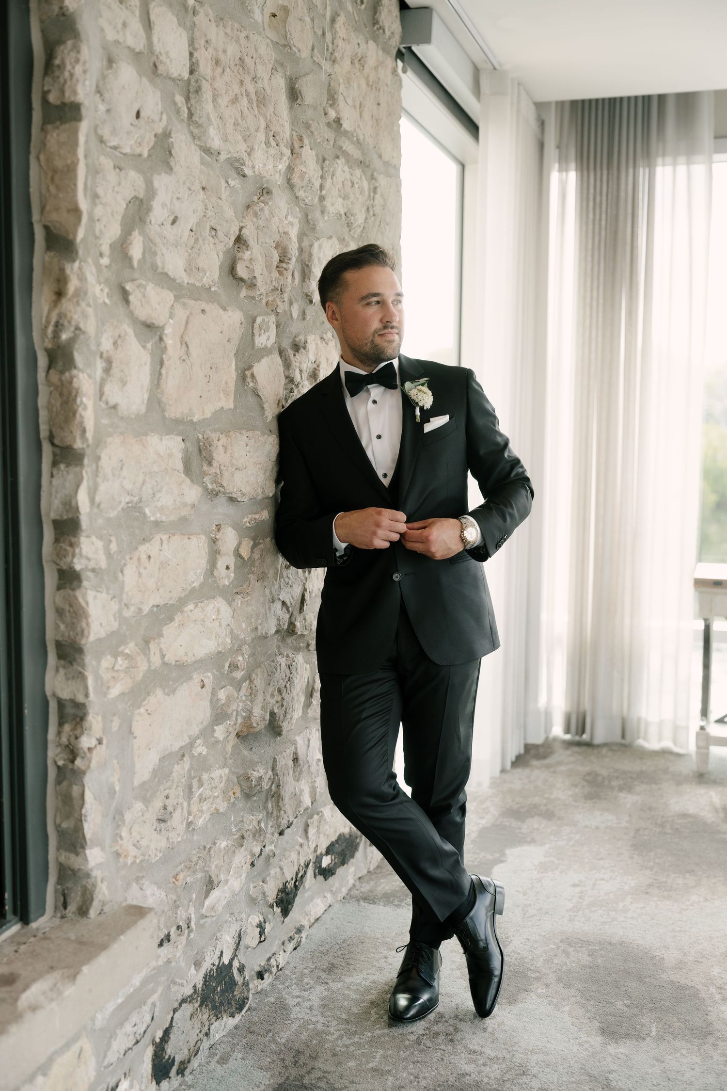 Dapper groom in black tuxedo with boutonniere leans against rustic stone wall by large windows at Cambridge Mill.