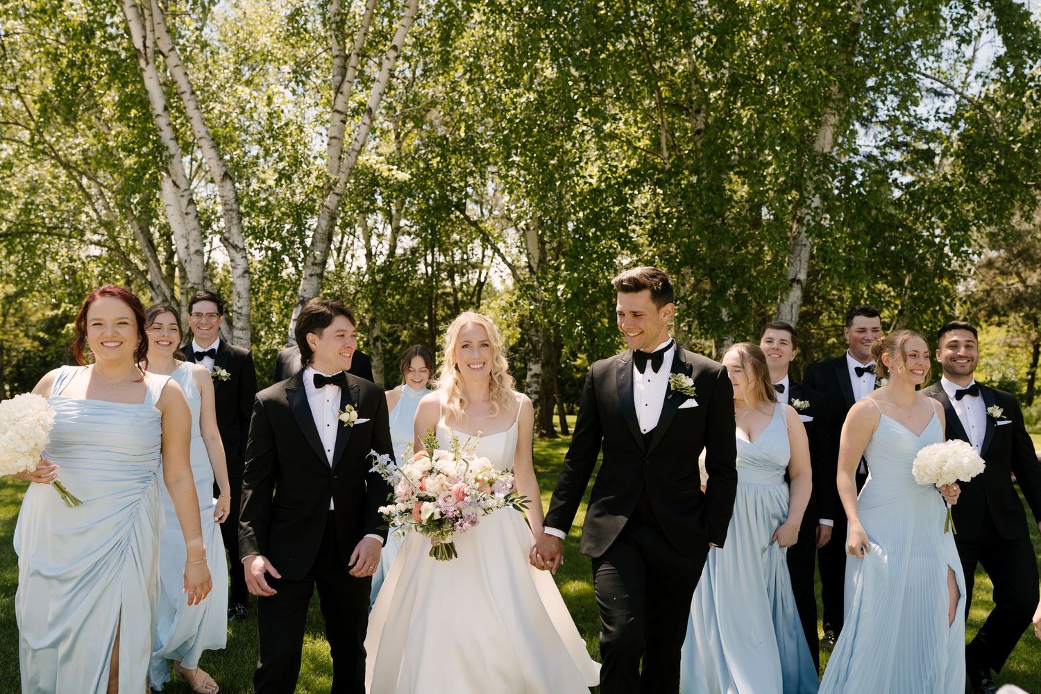 Wedding party walks together outdoors, bridesmaids in blue dresses and groomsmen in black tuxedos.