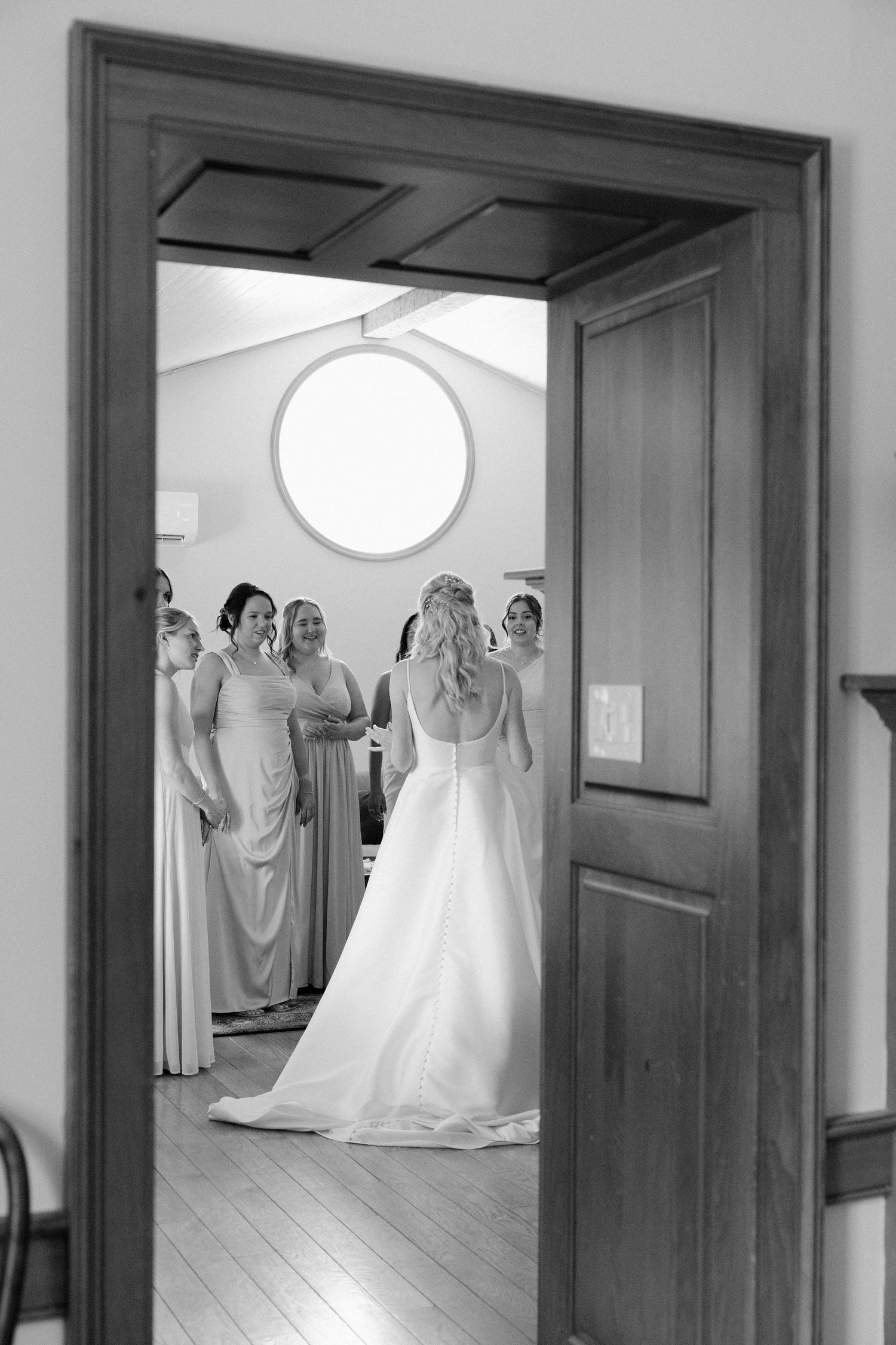Bride in white gown stands with bridesmaids viewed through an open wooden doorway at Roseville Estates.