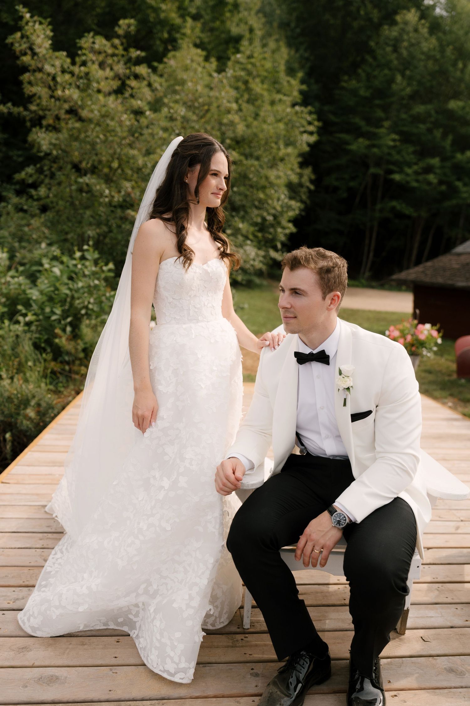 Elegant bride in lace gown and groom in white tuxedo jacket pose together on a wooden deck surrounded by lush greenery.