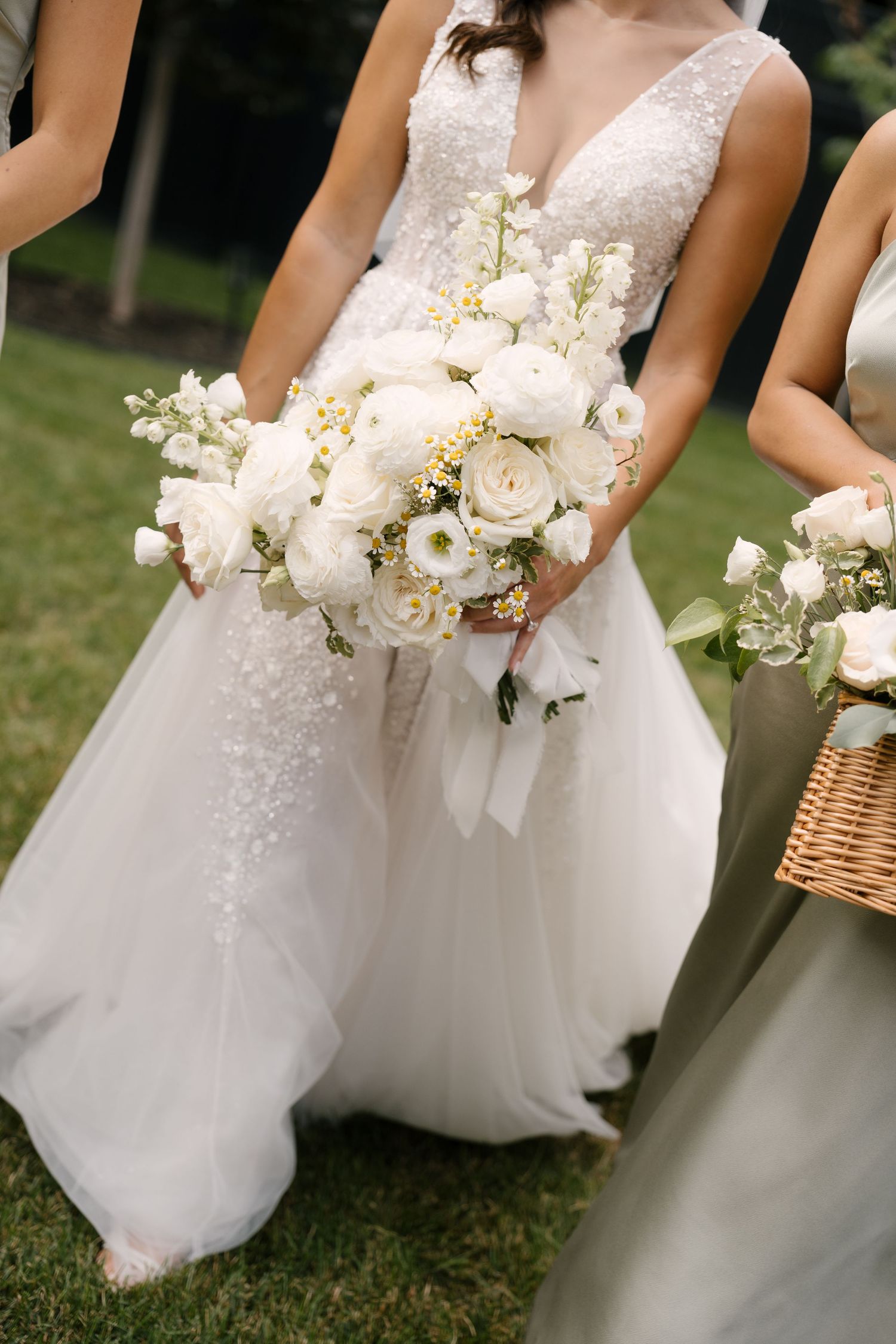 Bride in beaded white gown holds large white floral bouquet with bridesmaids in sage dresses outdoors.