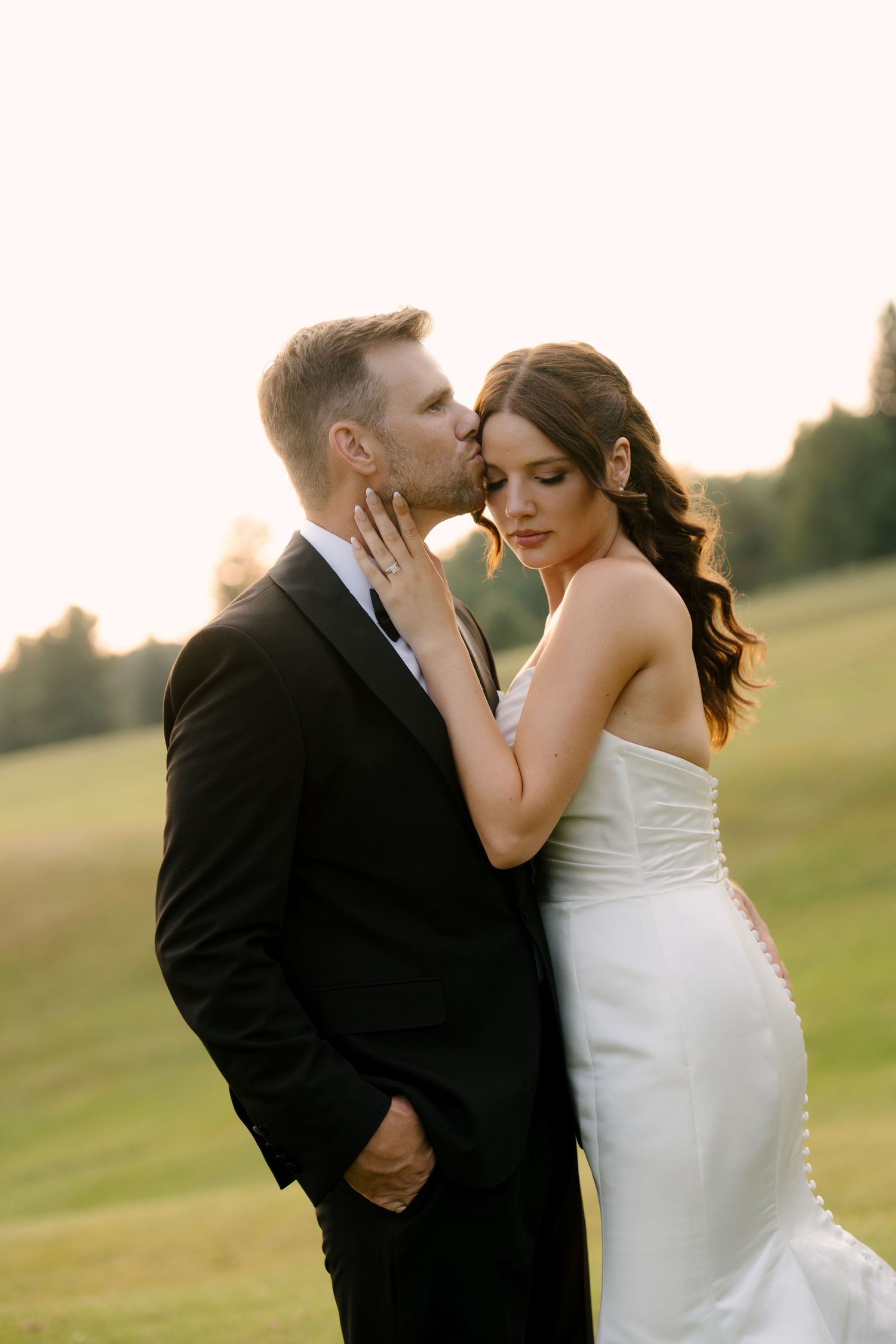 Groom kissing bride on the forehead at golden hour on a lush green golf course.