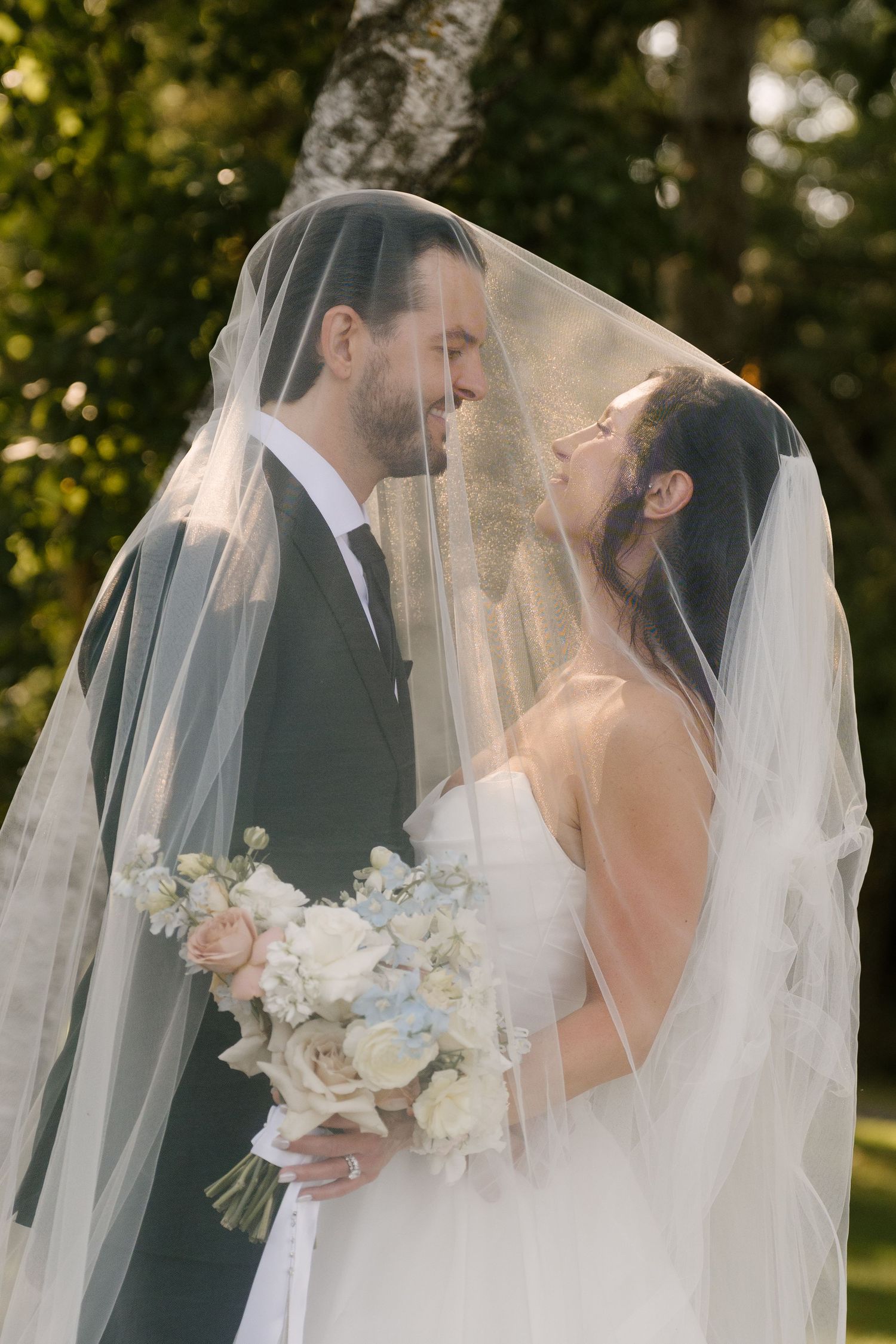 Bride and groom share a romantic moment under a wedding veil, bride holding a soft blue and white bouquet outdoors.