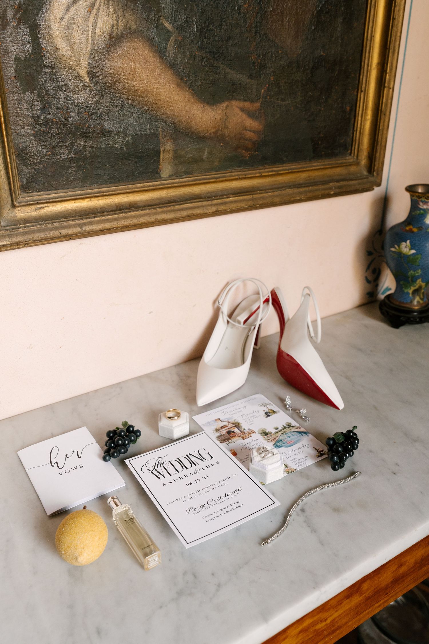 Flat lay of bridal accessories including red-soled white heels, wedding program, perfume, and jewelry on marble surface.