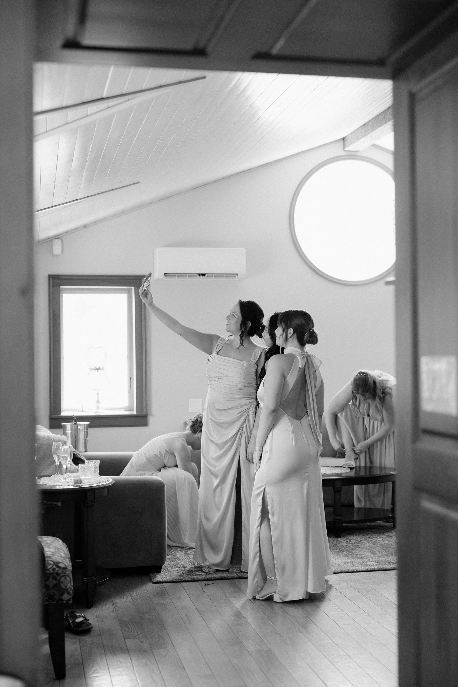 Bridesmaids in elegant gowns helping bride prepare in charming attic bridal suite, black and white photo.