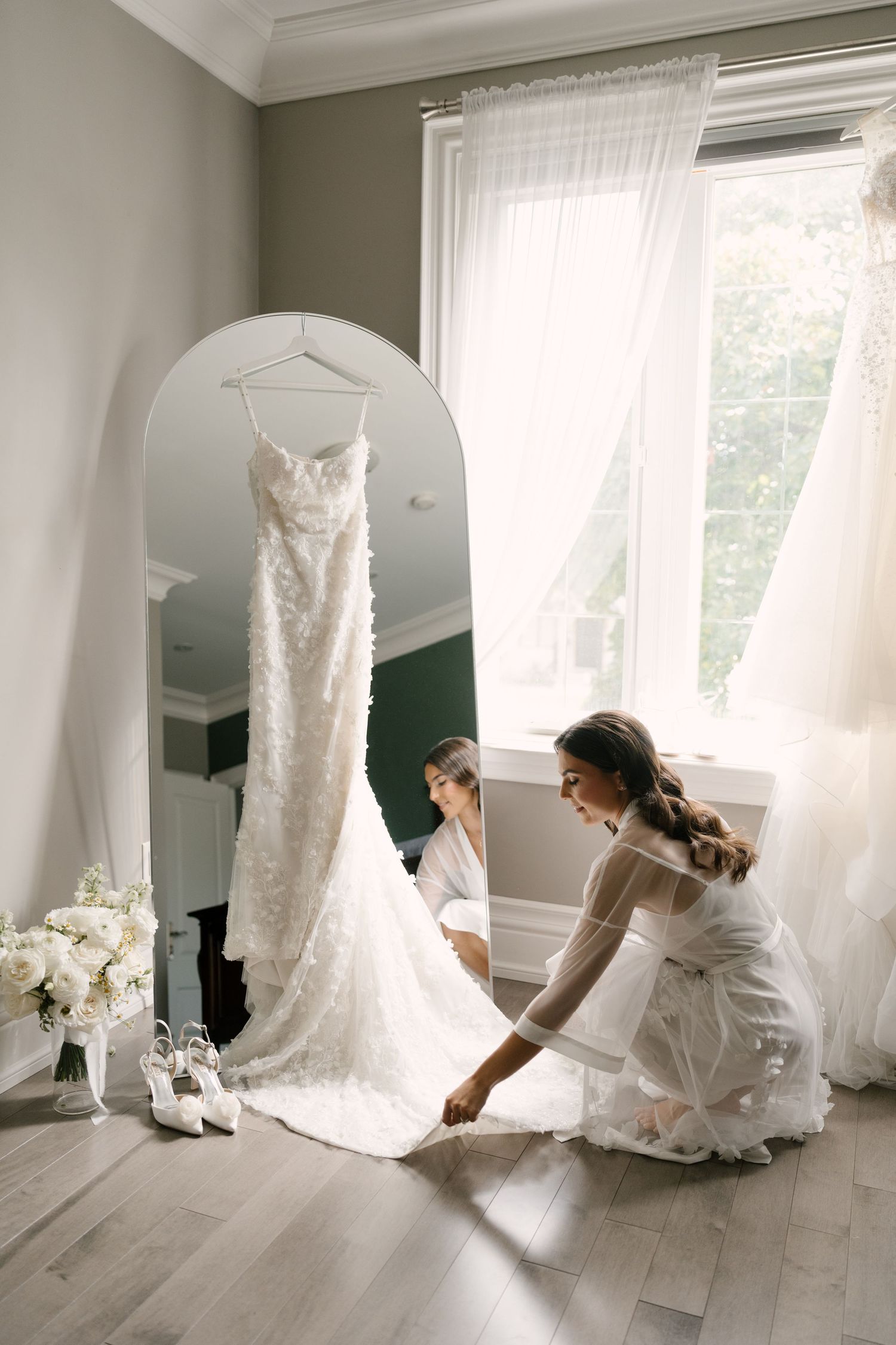 Bride in white lace robe adjusting her wedding gown train near a mirror and window in a bright, elegant room.