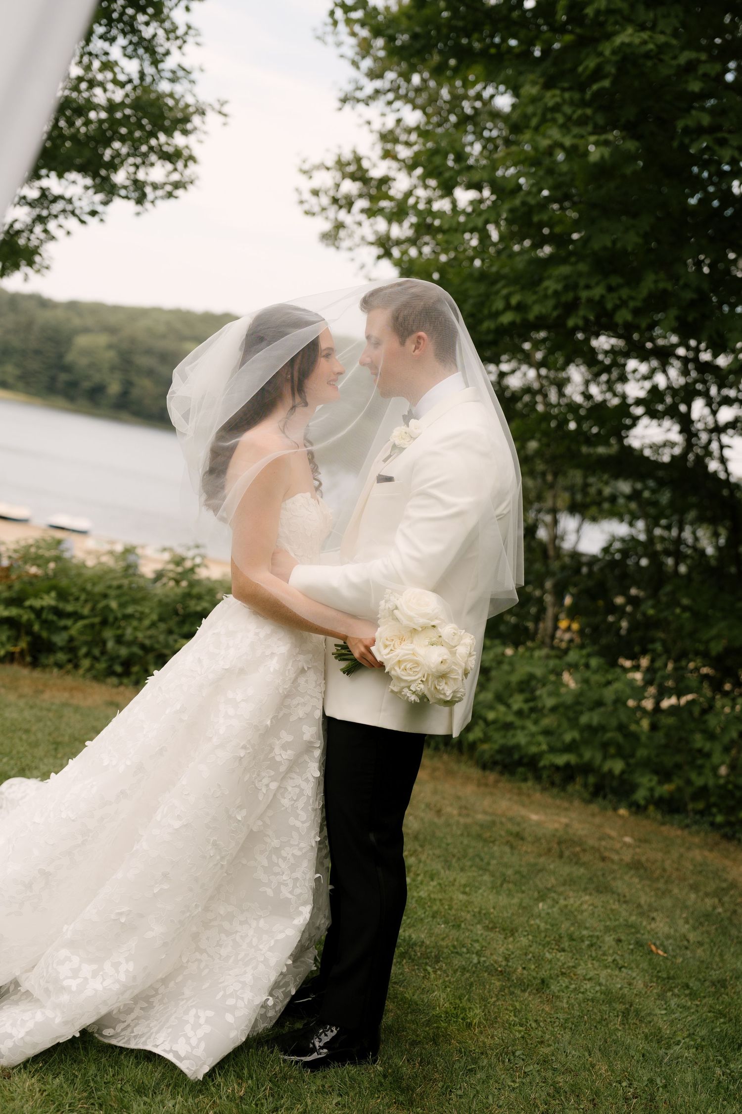 Bride and groom share romantic moment under veil outdoors by lake, bride in lace gown, groom in white tuxedo jacket.