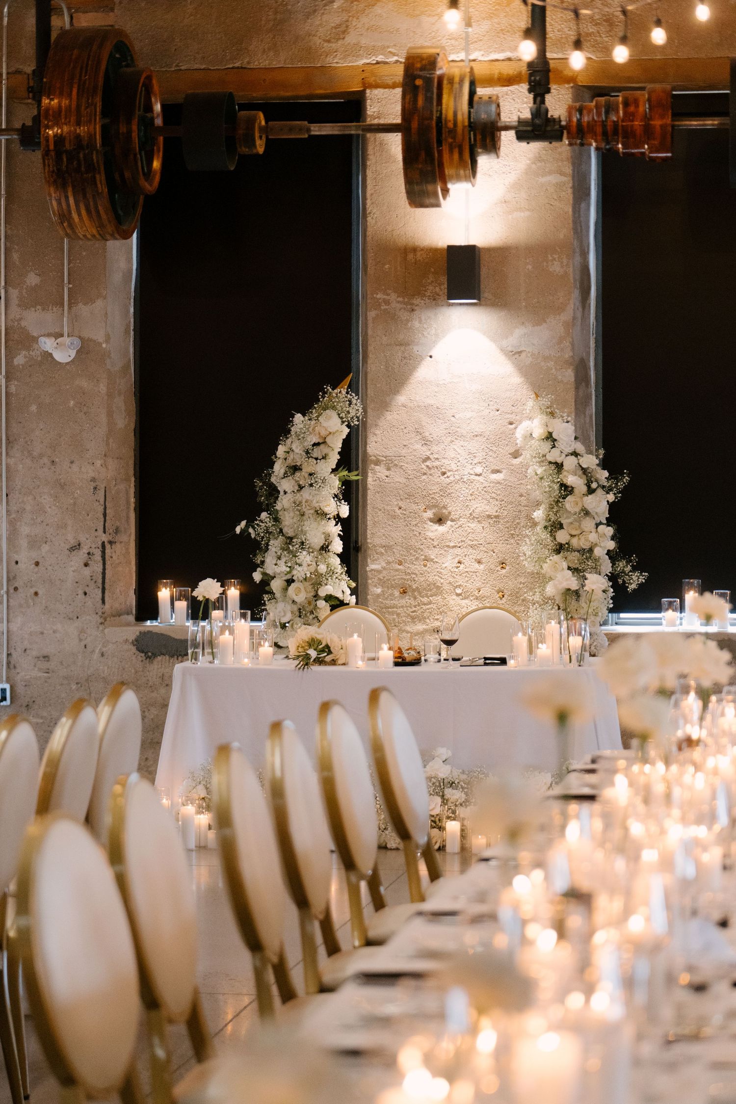 Elegant wedding reception with white floral arch, candlelit tables, gold chairs, and string lights in an industrial venue.