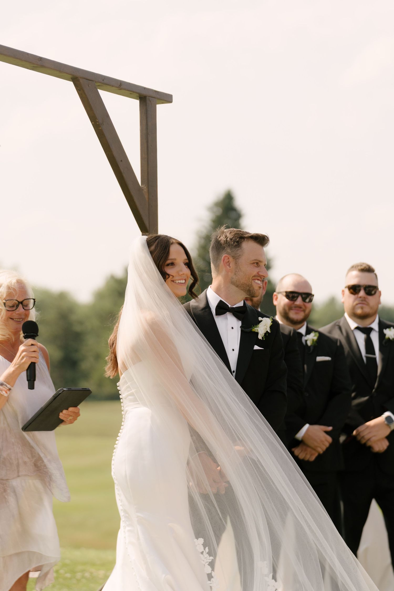 Bride and groom laugh joyfully at outdoor wedding ceremony beneath a wooden arch with groomsmen behind them.