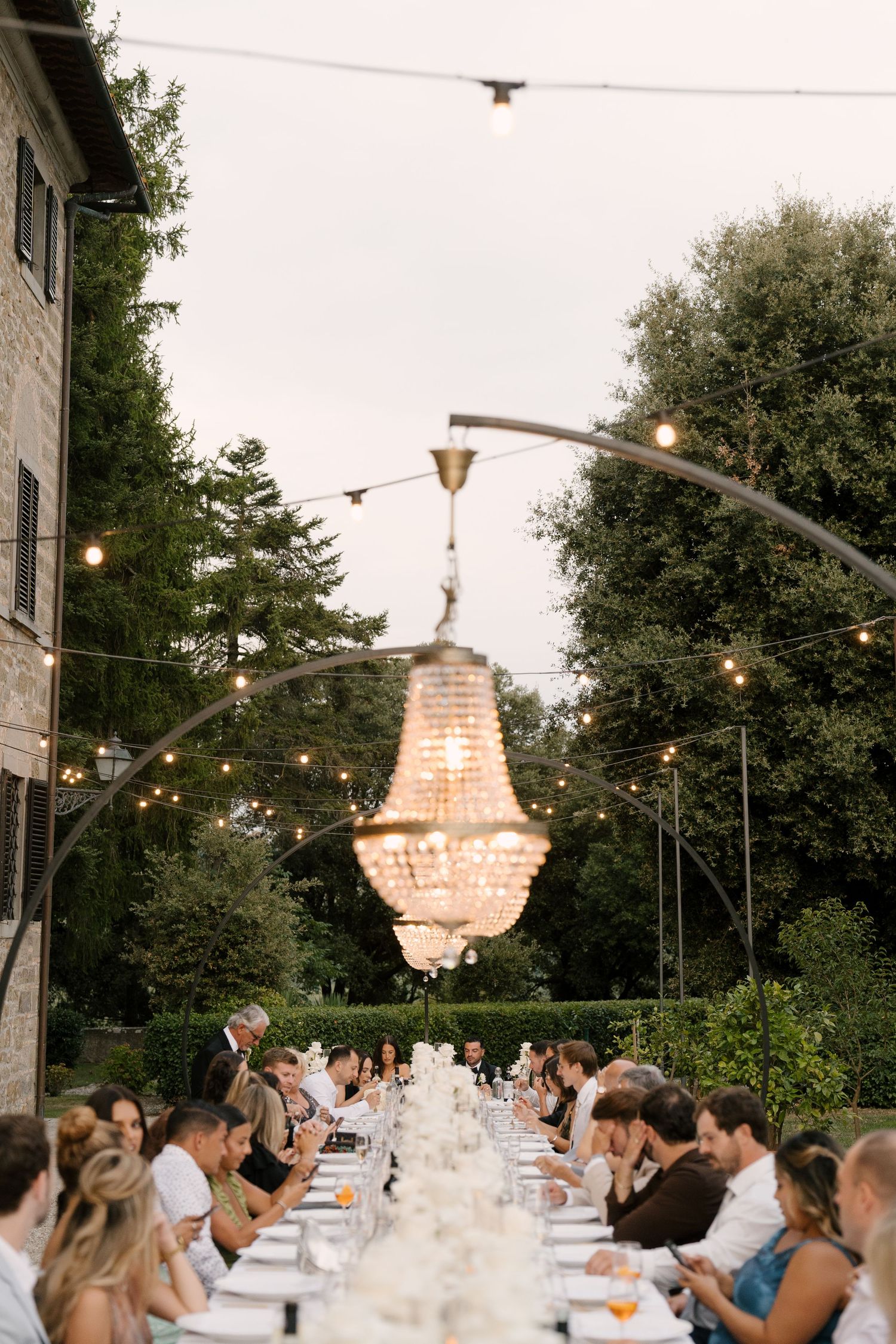 Guests dining at long outdoor Tuscan wedding reception table beneath glowing crystal chandelier at dusk.