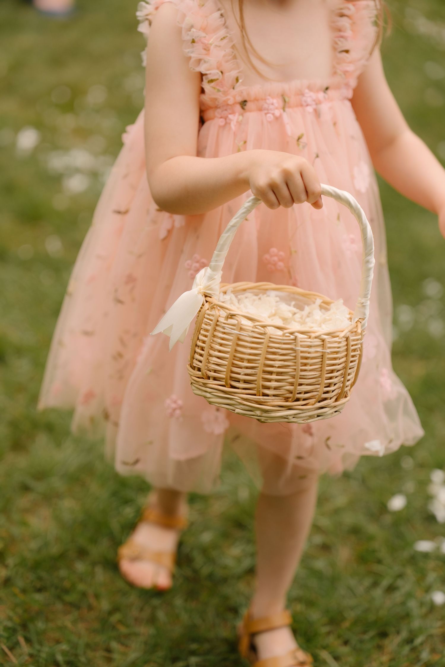 Young flower girl in a pink tulle dress carries a wicker basket during an outdoor wedding ceremony.