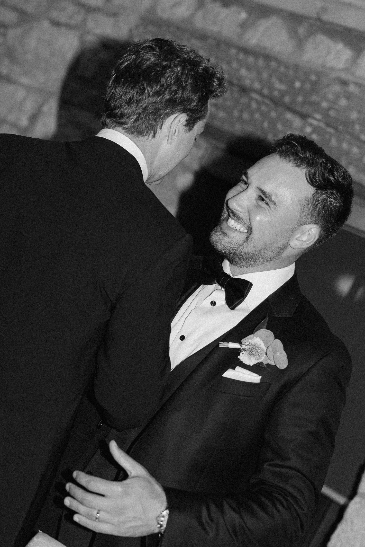 Joyful groom in tuxedo with boutonniere embraces guest while laughing at Cambridge Mill wedding reception.
