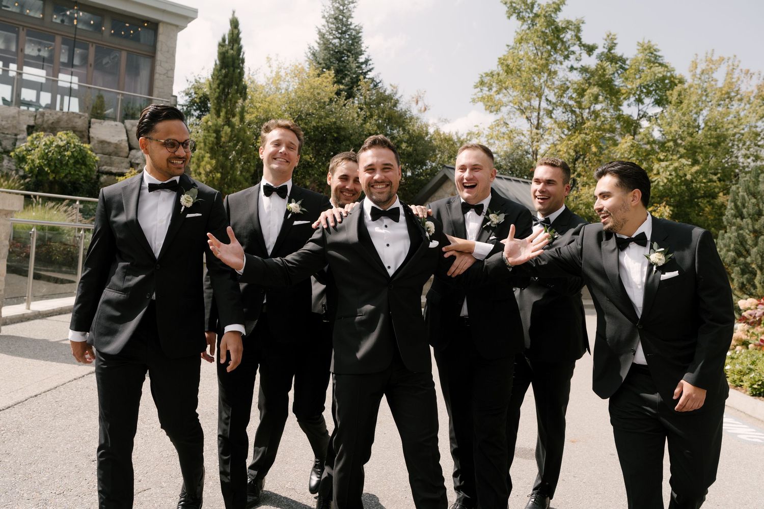 Groom and groomsmen in black tuxedos with bow ties laughing together outdoors at a wedding celebration at Cambridge Mill.