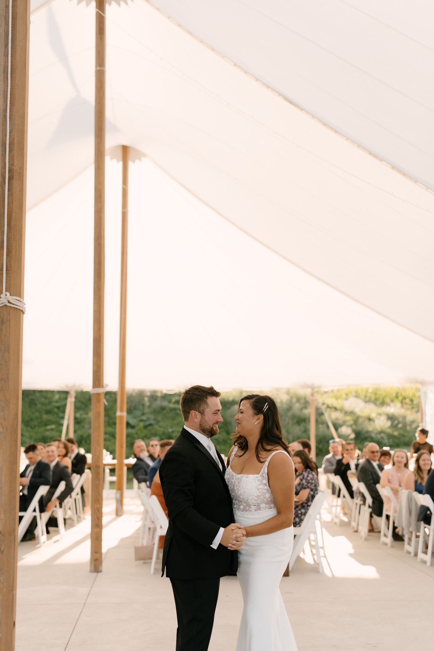 Bride and groom share a romantic first dance under a white tent during their outdoor wedding ceremony with guests seated behind them.