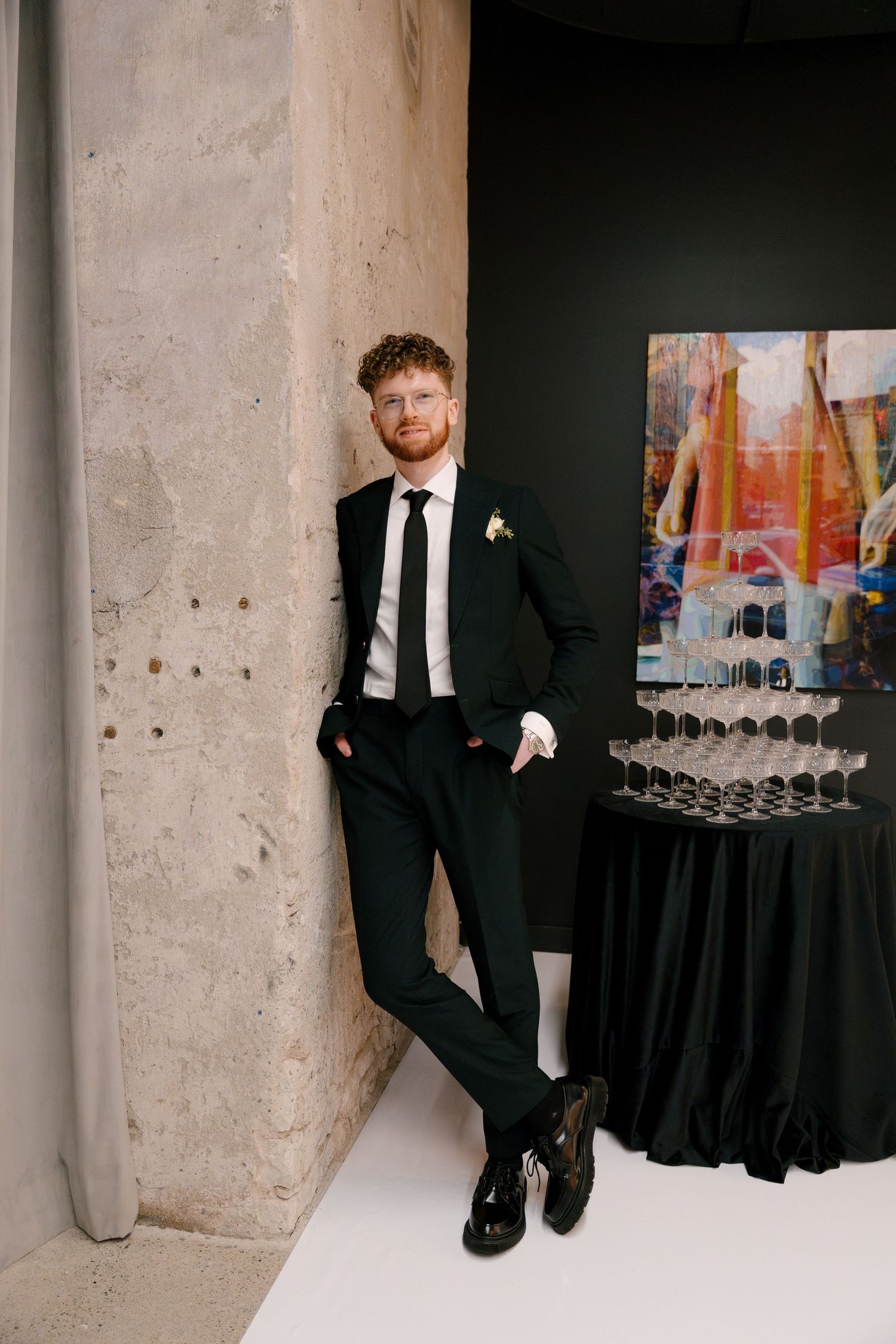 Groom in black suit with white boutonniere leans against concrete wall near champagne tower at wedding reception.
