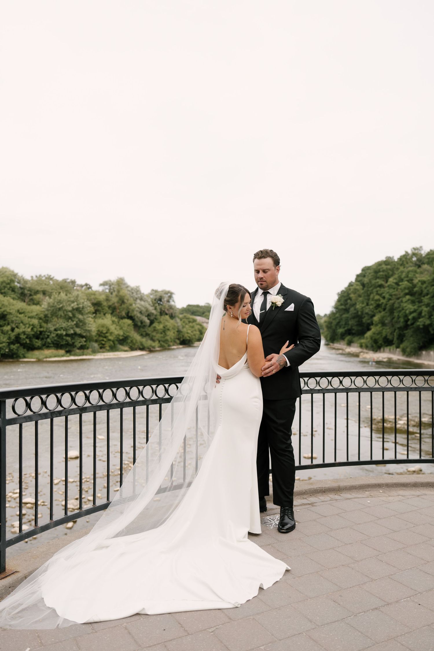 Bride and groom share romantic moment on bridge overlooking river in elegant wedding attire.