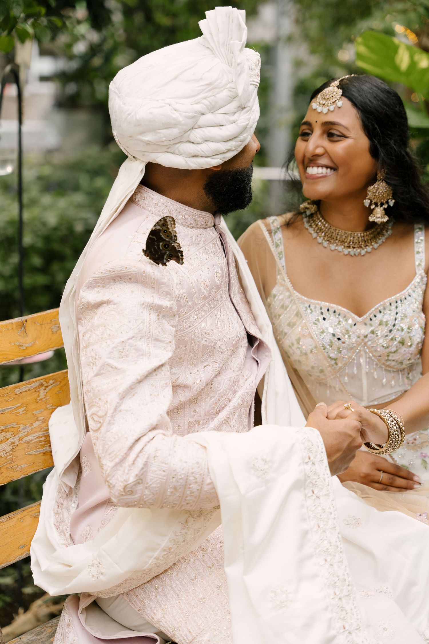 Indian couple in traditional wedding attire sit on bench laughing with butterfly on groom.