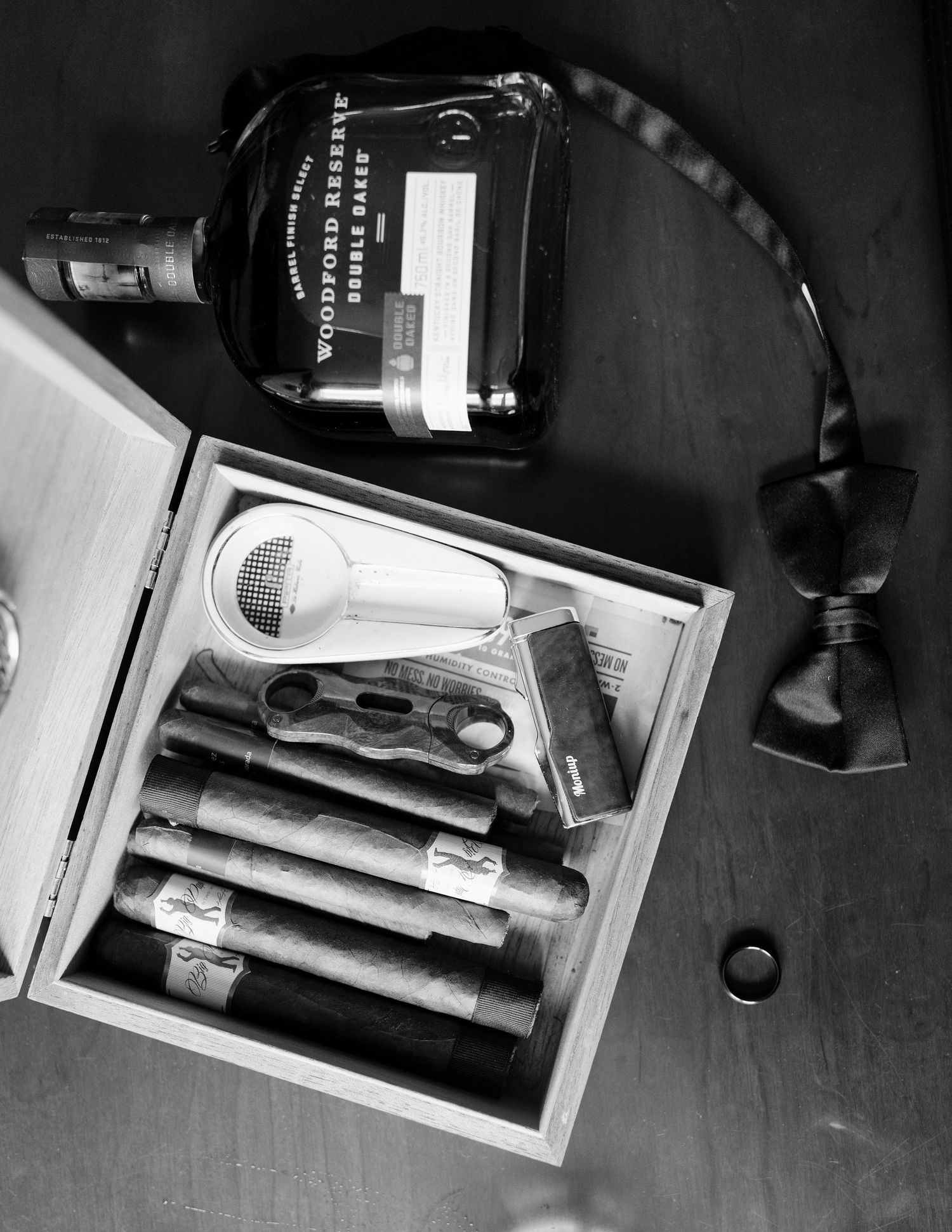 Black and white flat lay of cigars, Woodford Reserve whiskey, and a bow tie for a groom.