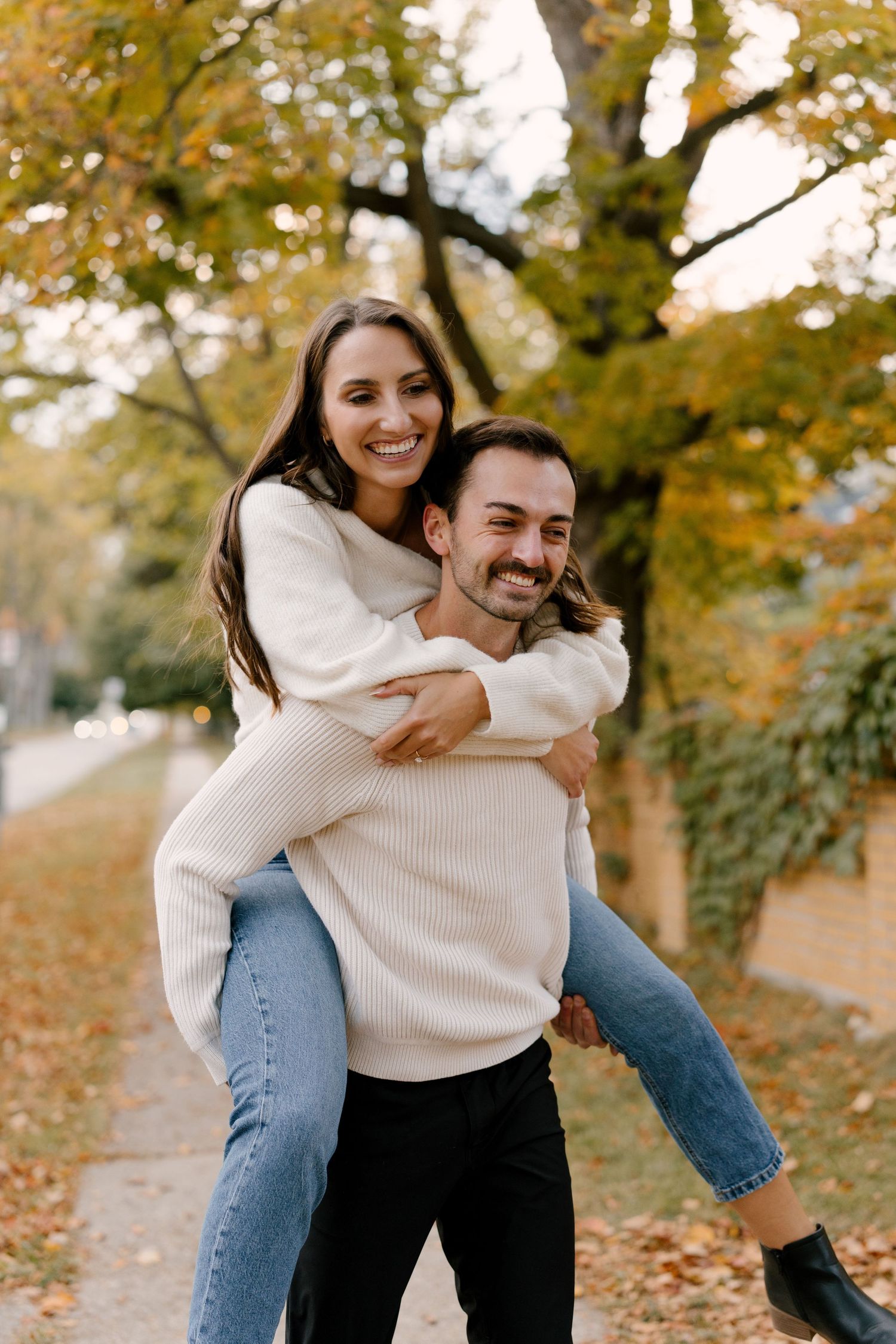 Man giving woman a piggyback ride on a tree-lined path with golden autumn foliage.