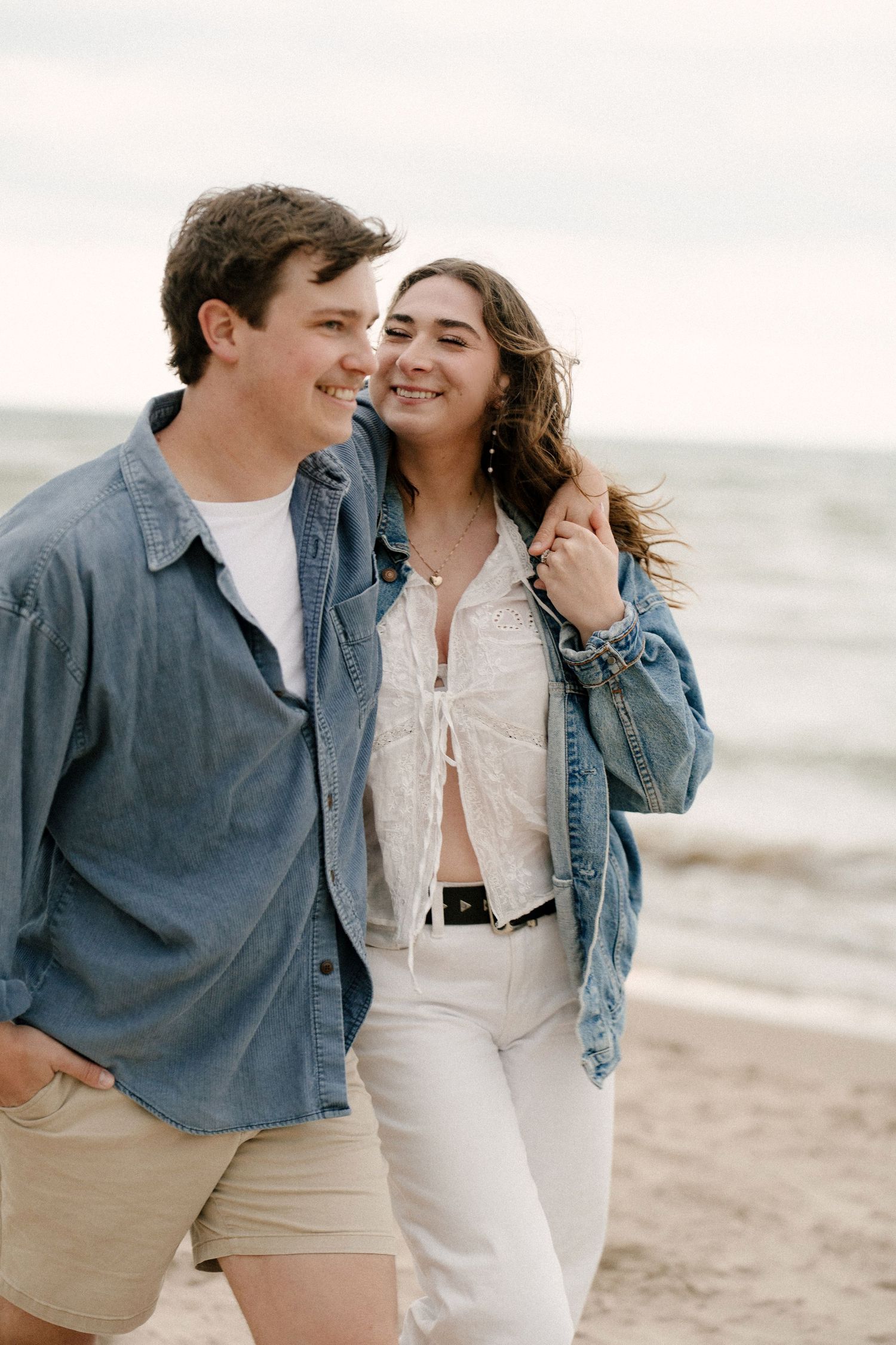 Smiling couple walking closely together on a beach, woman wearing a denim jacket over a white outfit.