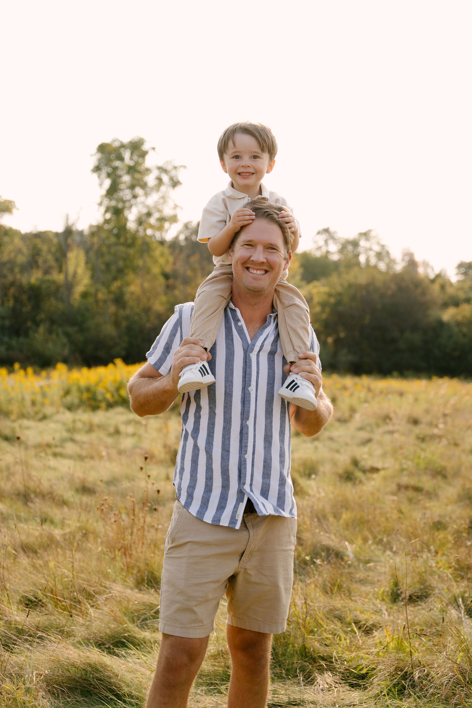 Smiling father carries young son on his shoulders in a sunny golden field with wildflowers and trees in background.