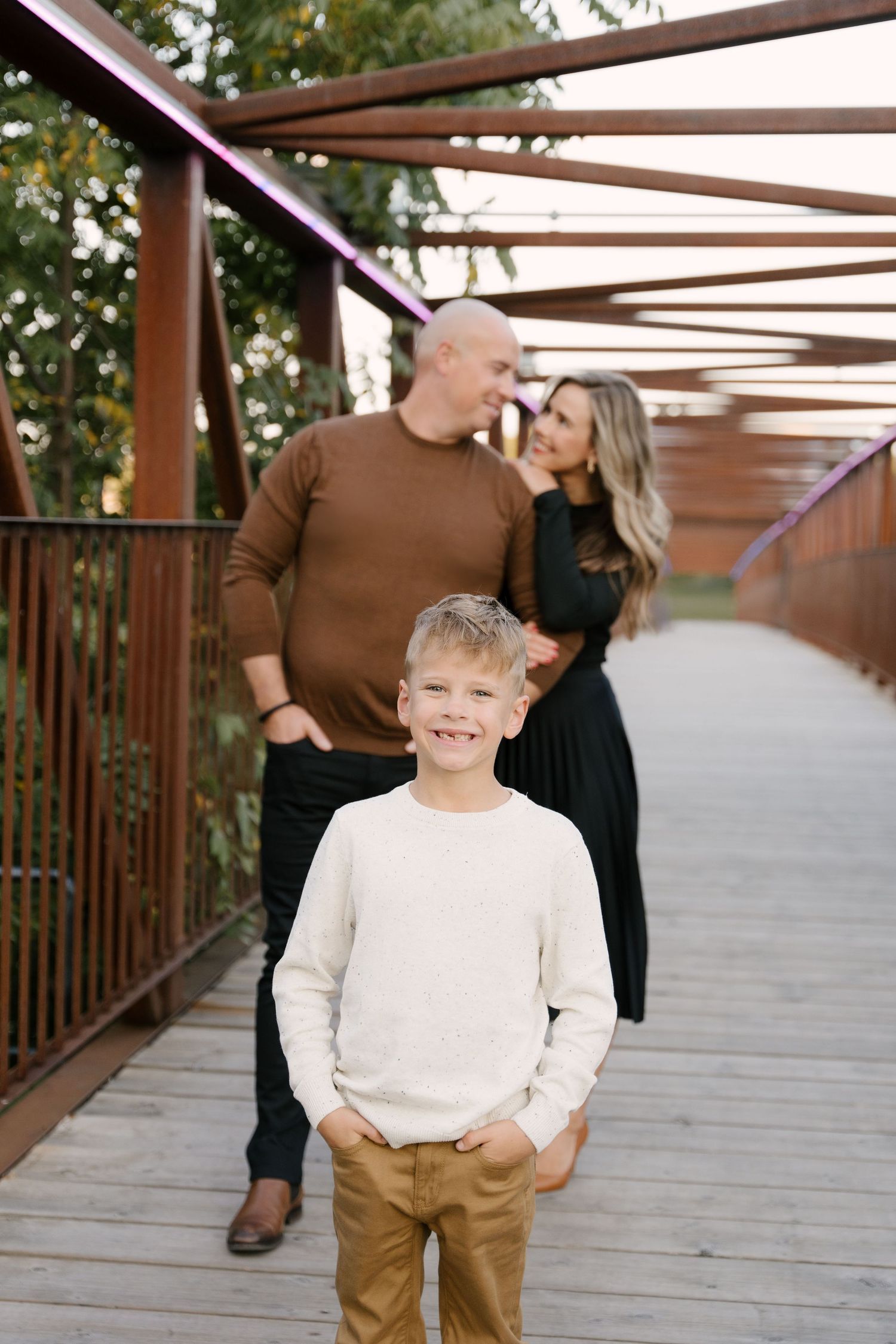 Young boy smiling at camera while parents share a romantic moment on bridge.