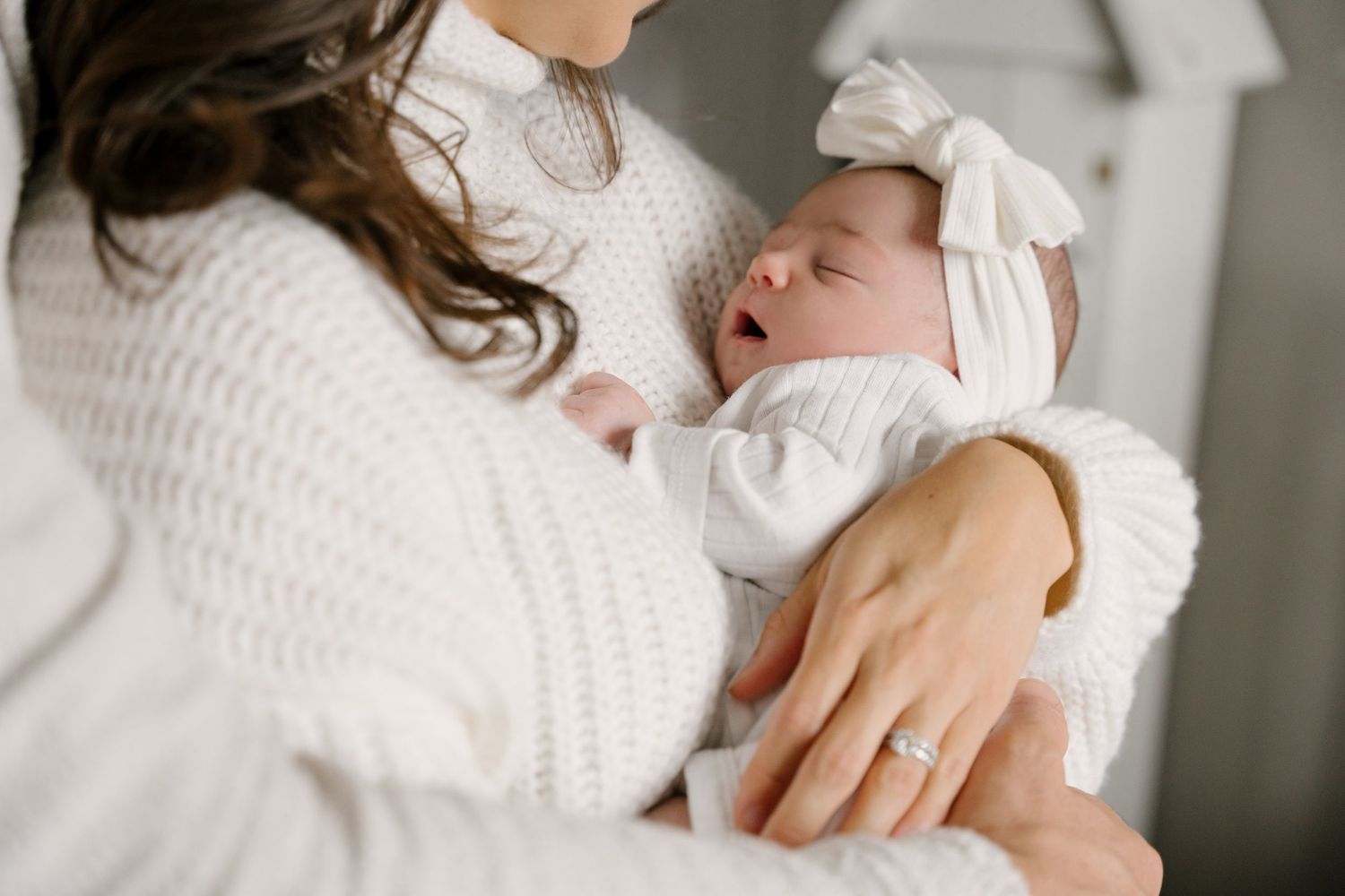 Mother in white knit sweater cradling sleeping newborn baby dressed in white with bow headband.