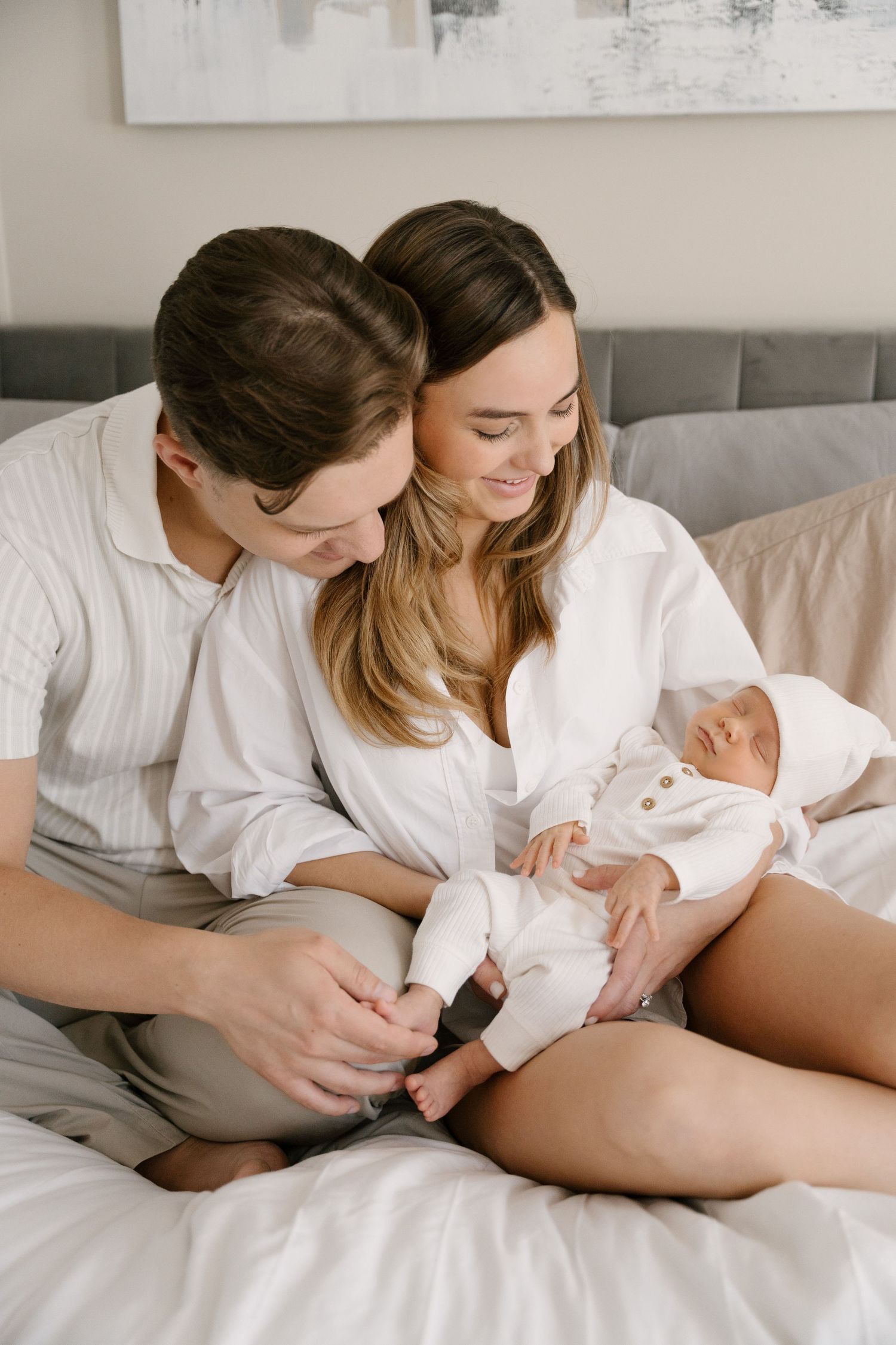 Young couple sitting on bed smiling at their newborn baby in white outfit.