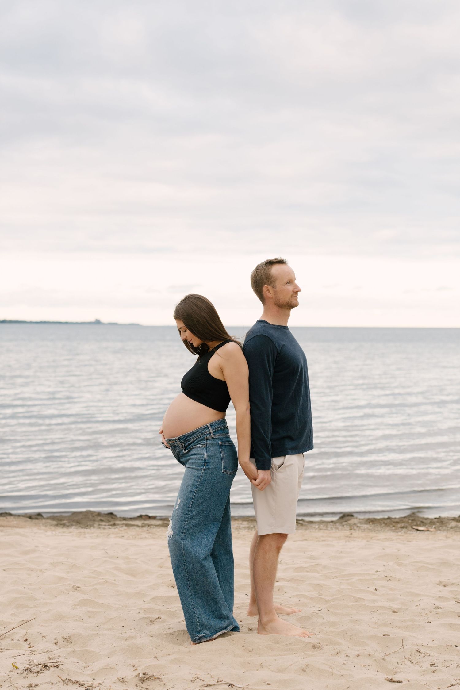 Pregnant couple stands back-to-back on sandy beach, woman holding baby bump while man gazes at the ocean.
