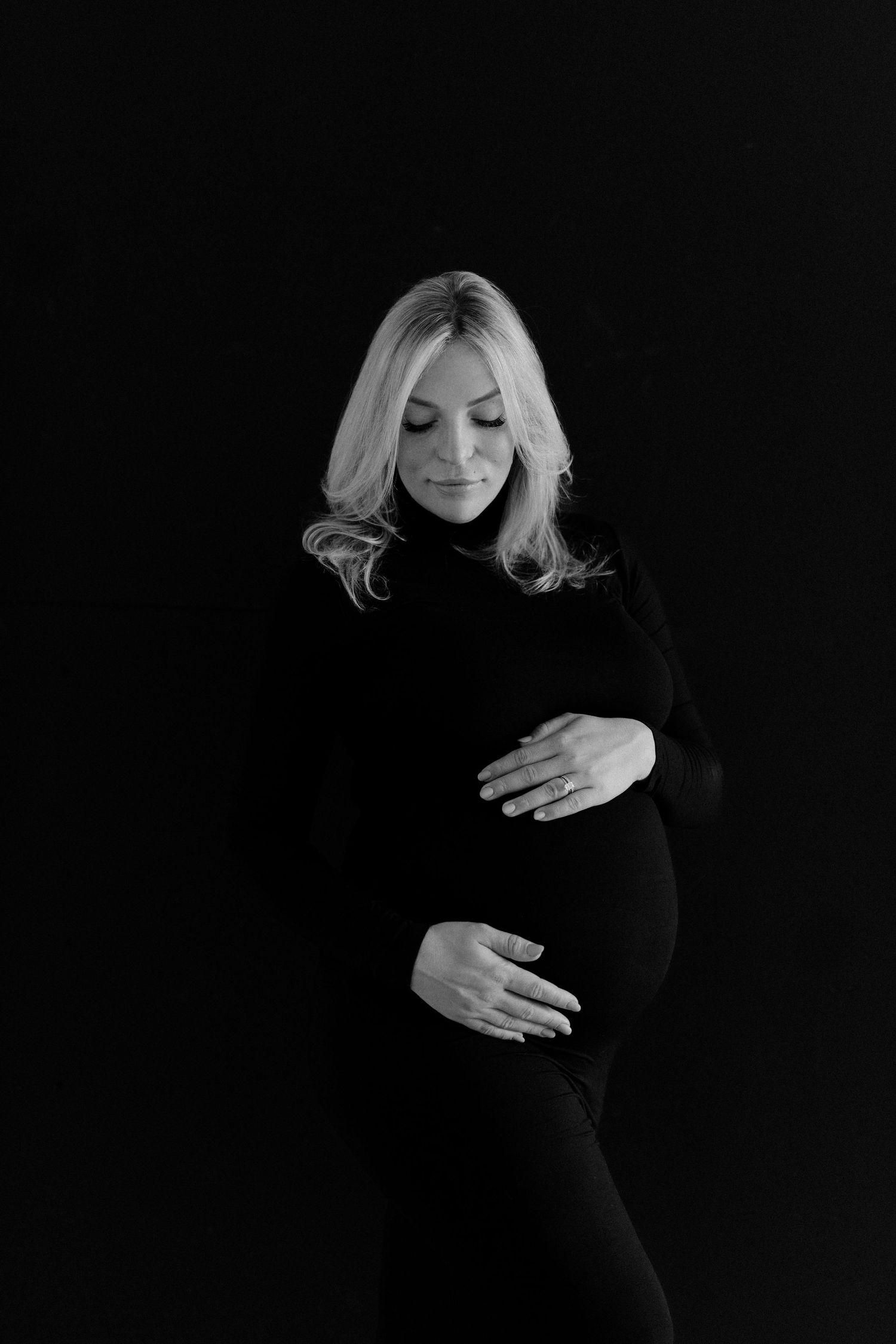 Black and white portrait of pregnant blonde woman in black outfit tenderly cradling her baby bump.