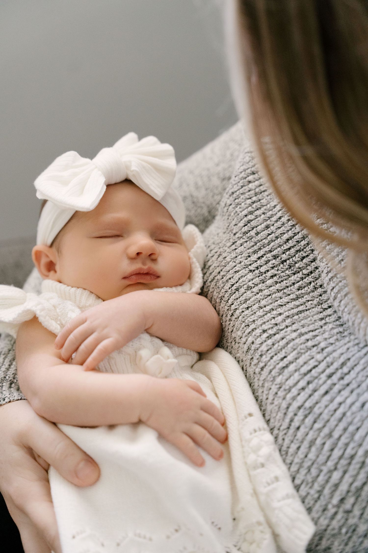 Peaceful newborn baby girl in white knit outfit and bow headband sleeping in mothers arms.