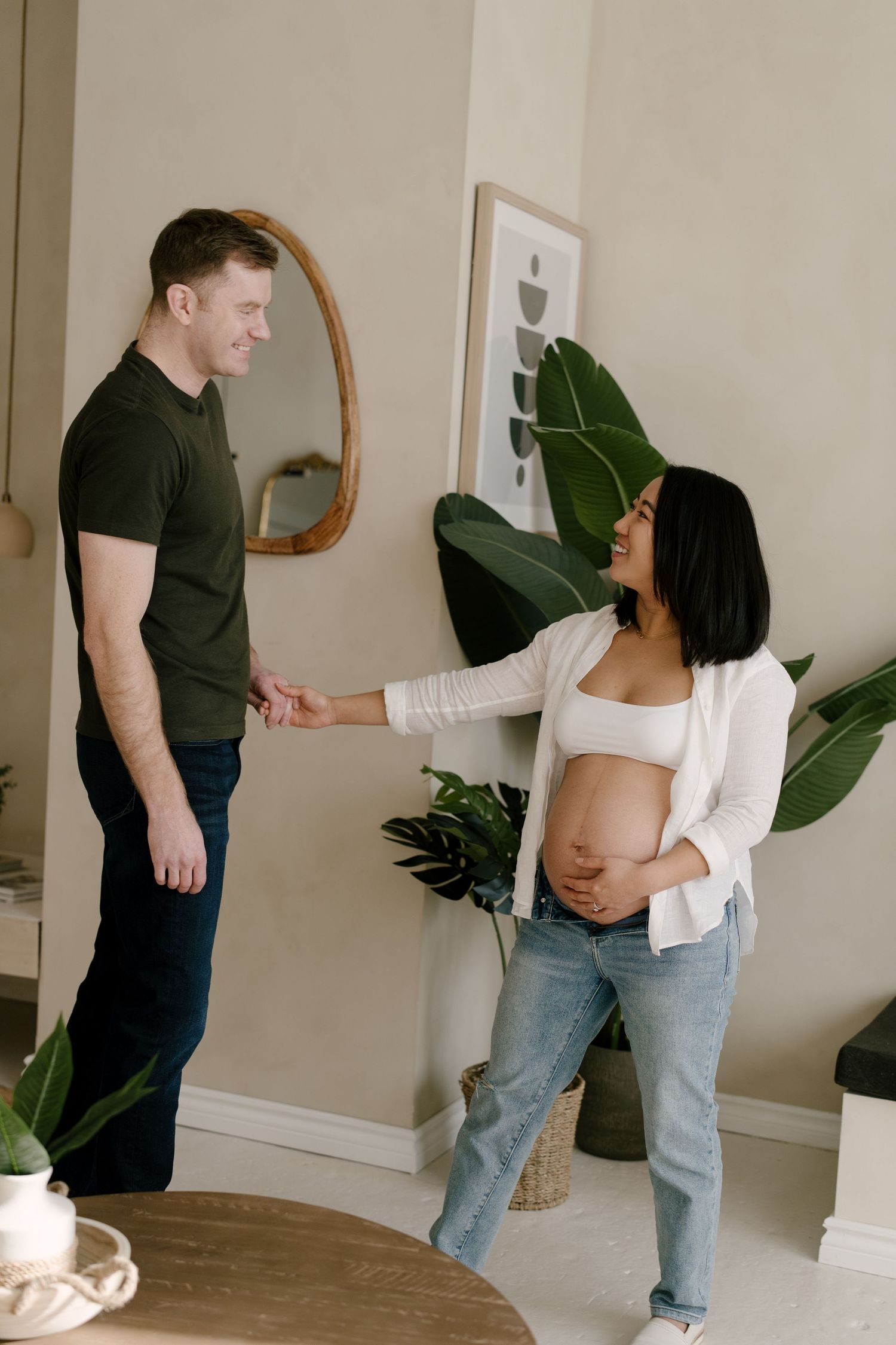 Smiling pregnant woman in white top and jeans holds hands with man in olive shirt in modern home.