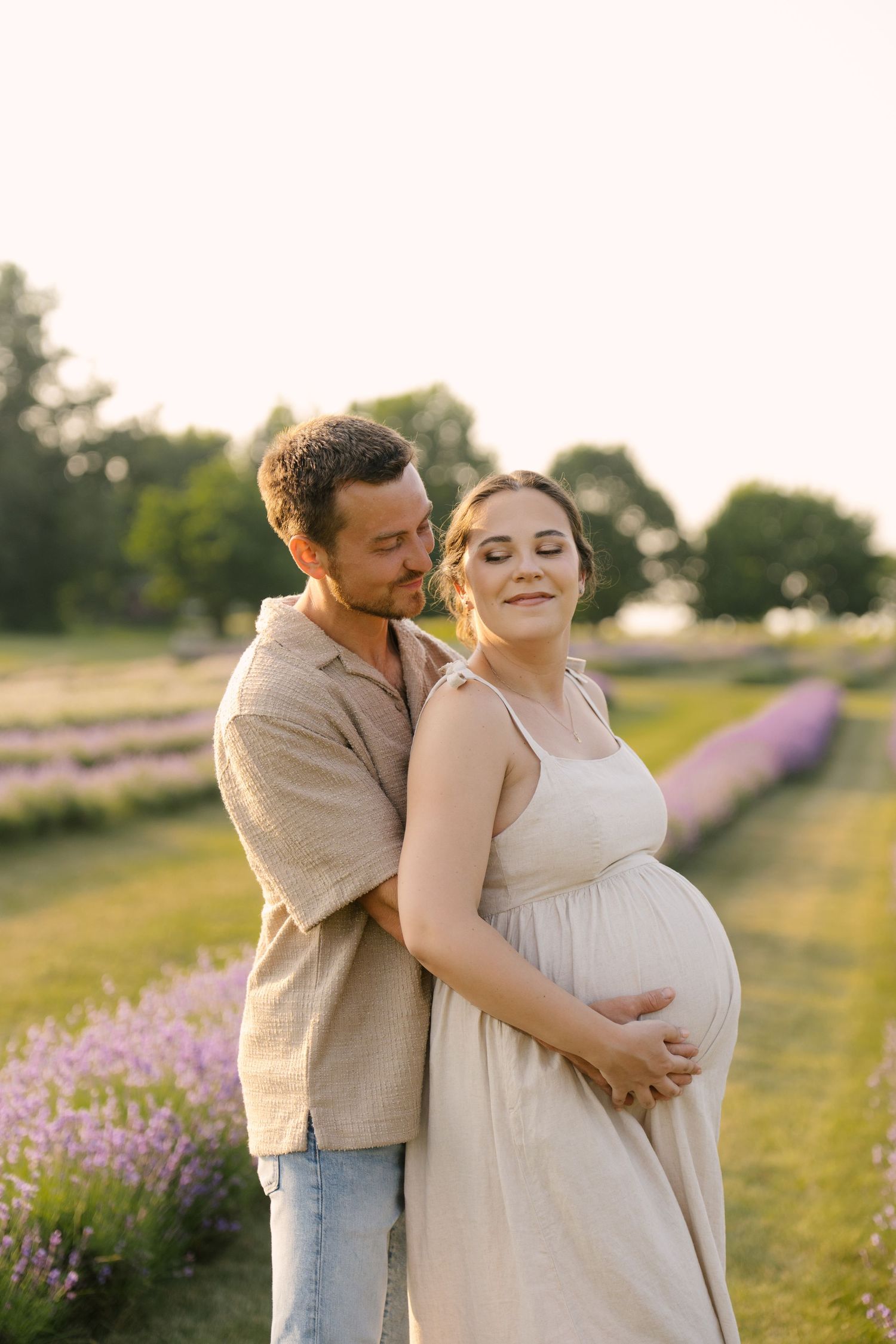 Couple embracing in lavender field at golden hour, man hugging pregnant woman from behind tenderly.