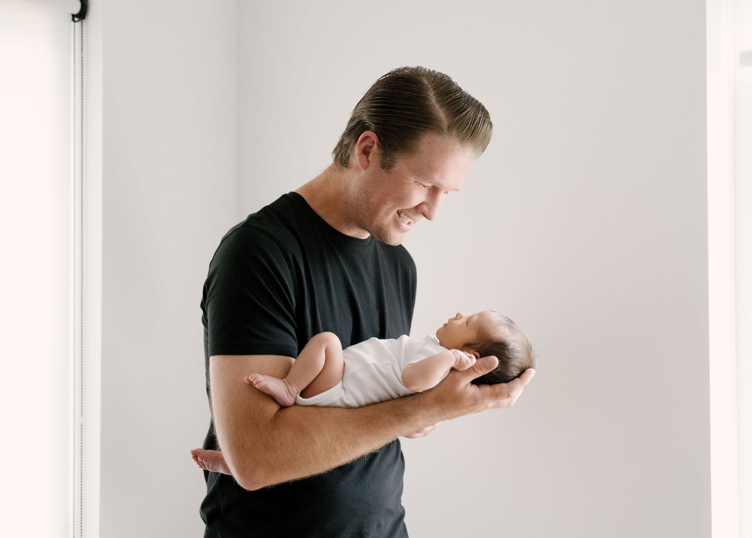 Smiling father in black t-shirt holding newborn baby near bright white window indoors.