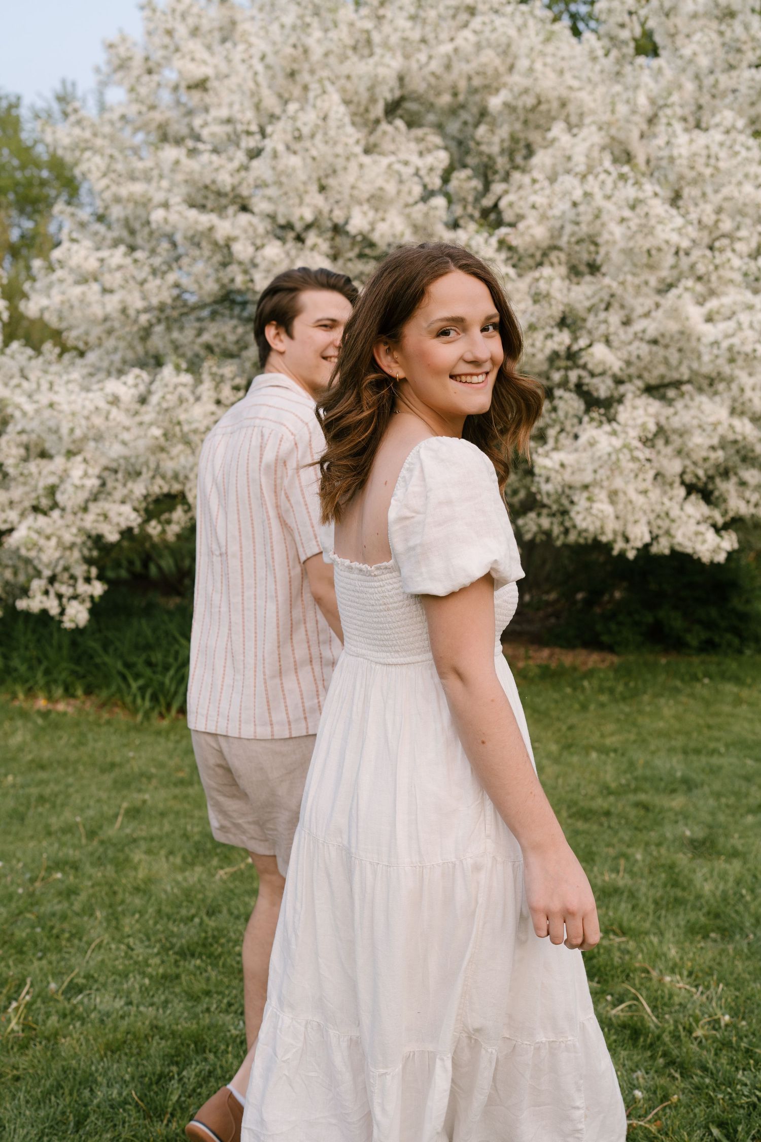 Smiling woman in white dress glancing back while walking with partner near a blooming white flowering tree.