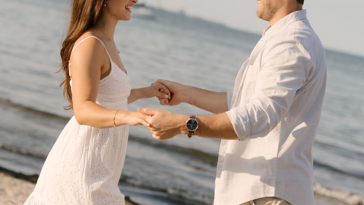 Couple holding hands on beach at sunset, woman in white dress smiling, man wearing a stylish watch.