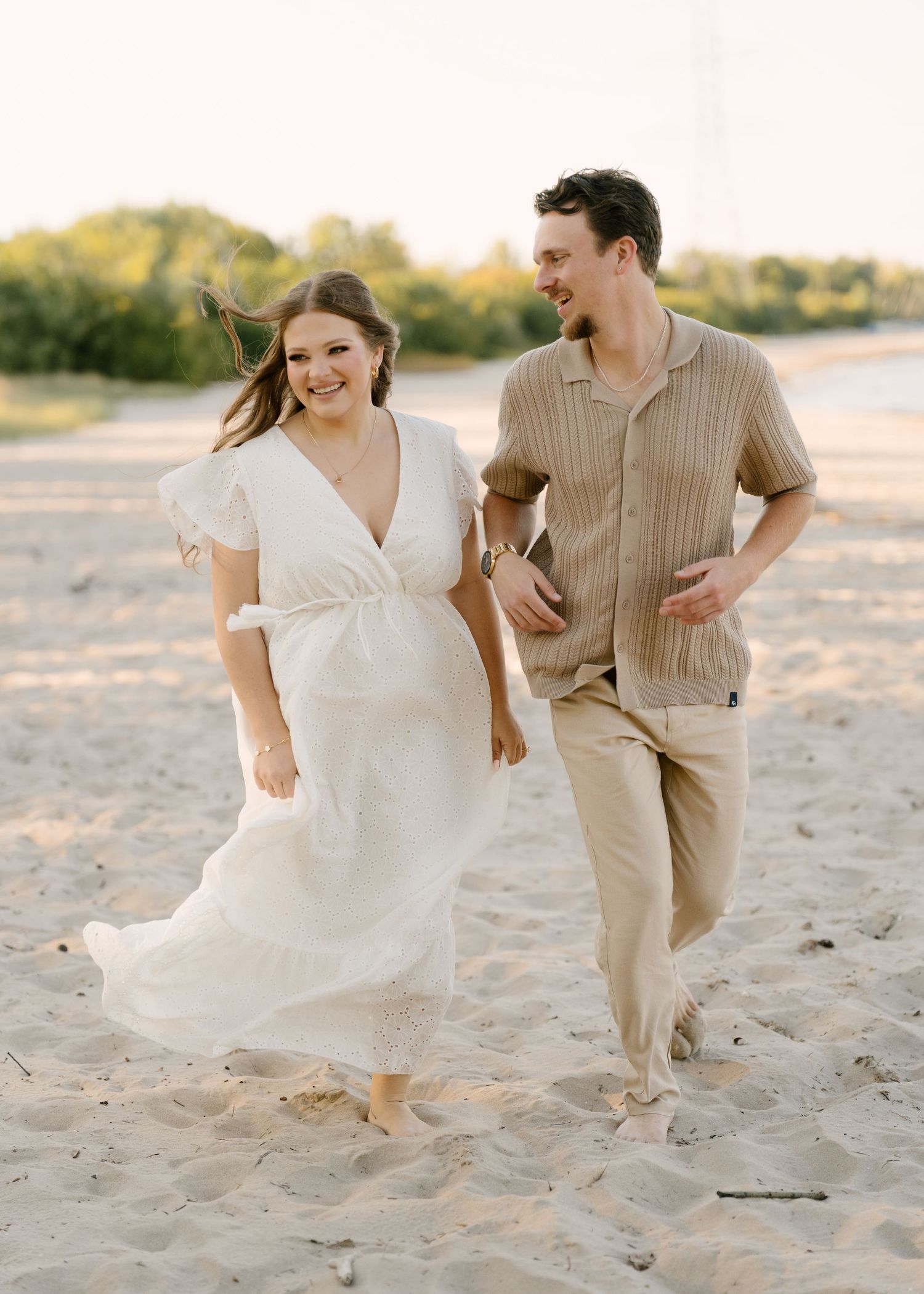Happy couple walking hand in hand on a sandy beach at sunset in coordinating neutral outfits.