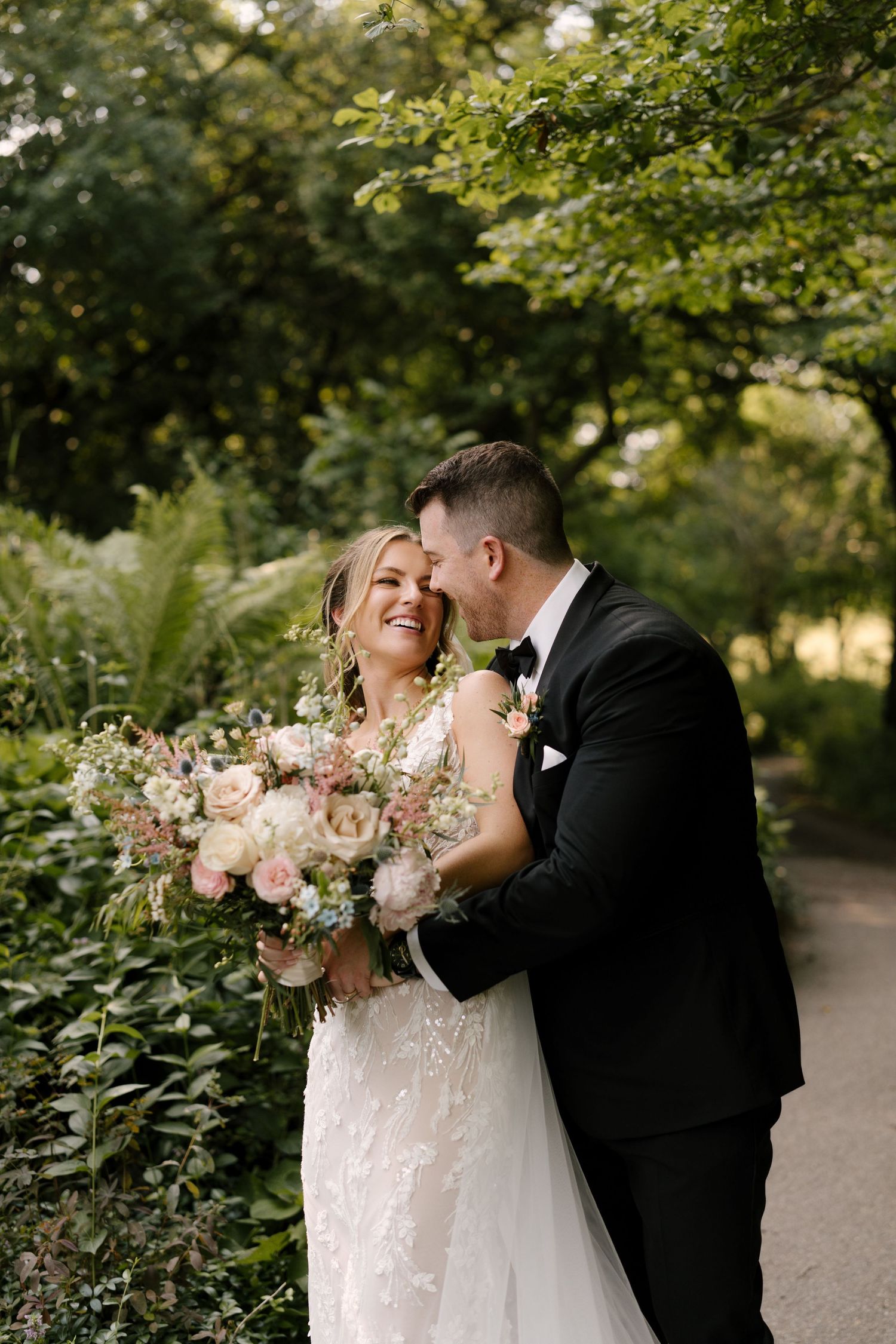 Groom whispers to laughing bride holding large pink and white bouquet in a sunlit garden setting.