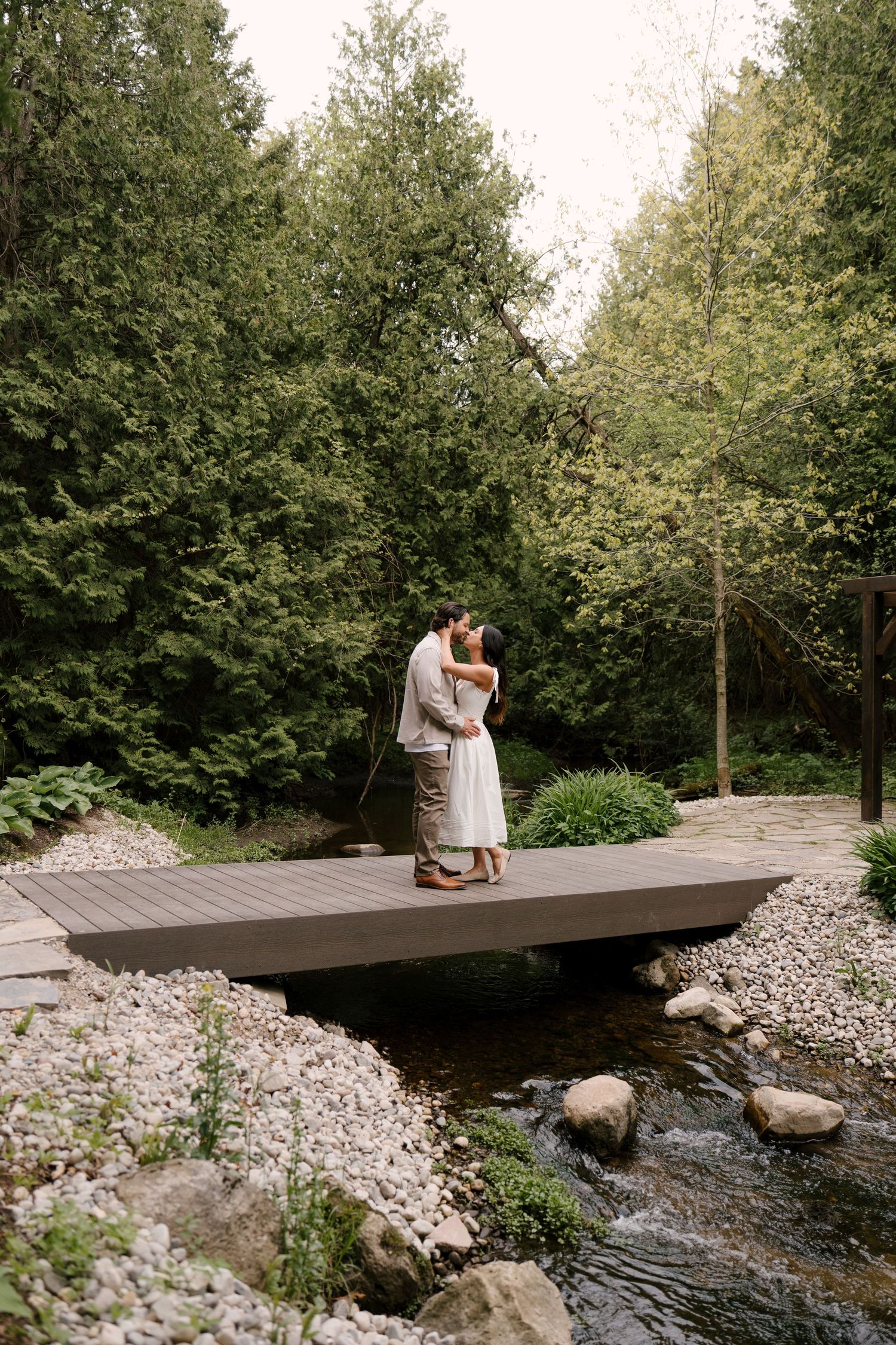Couple sharing a kiss on a wooden bridge over a stream in lush green forest.