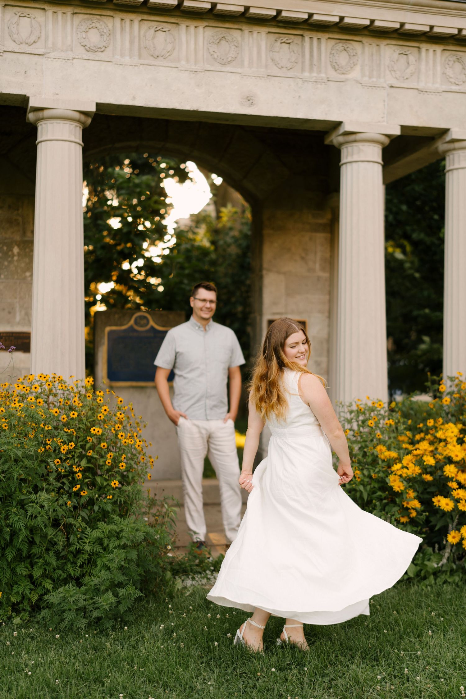 Woman in white dress twirling near a columned pavilion with yellow flowers as man watches.
