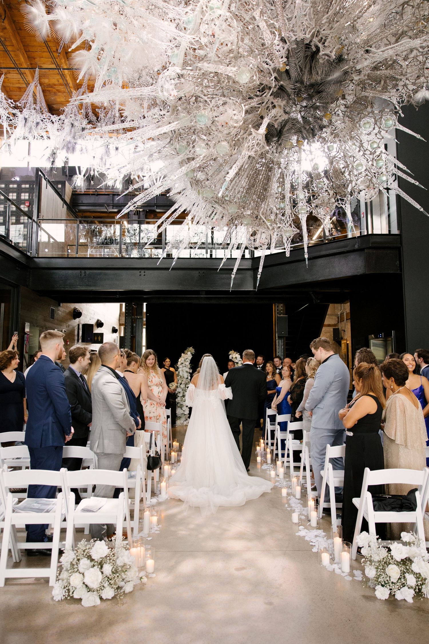 Wedding ceremony aisle lined with candles and white florals leading to altar beneath dramatic white sculptural chandelier at Tapestry Hall.