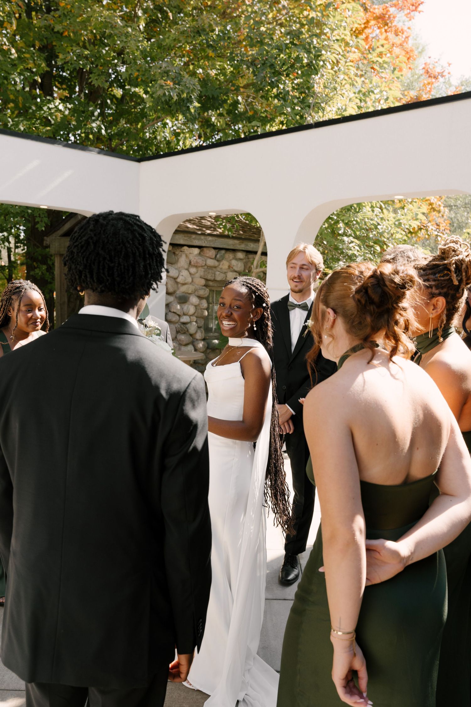 Interracial wedding couple greeting guests outdoors under a white arch surrounded by fall foliage at Wren House.