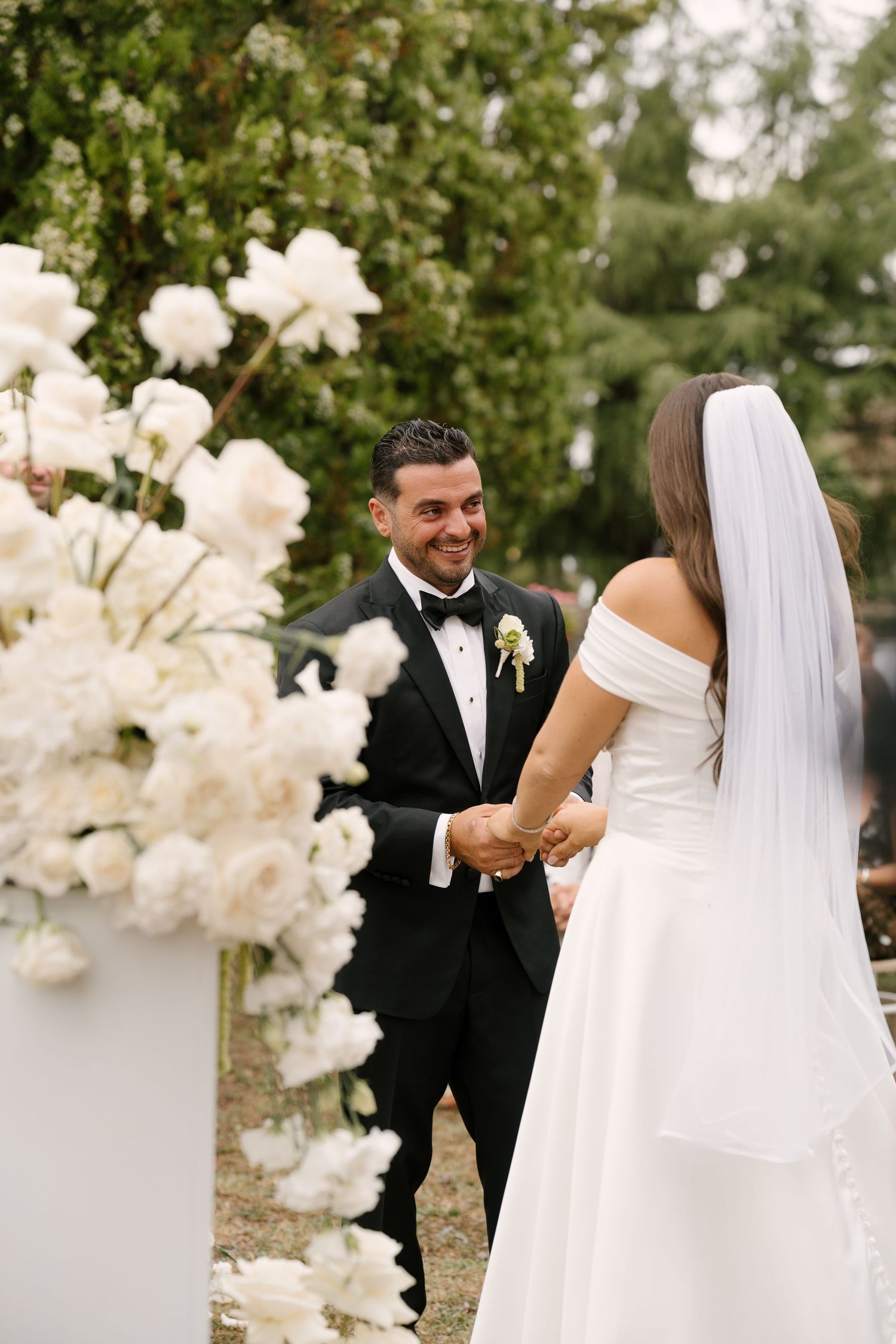 Groom in black tuxedo smiles at bride in white off-shoulder gown during outdoor wedding ceremony with white floral arrangements.