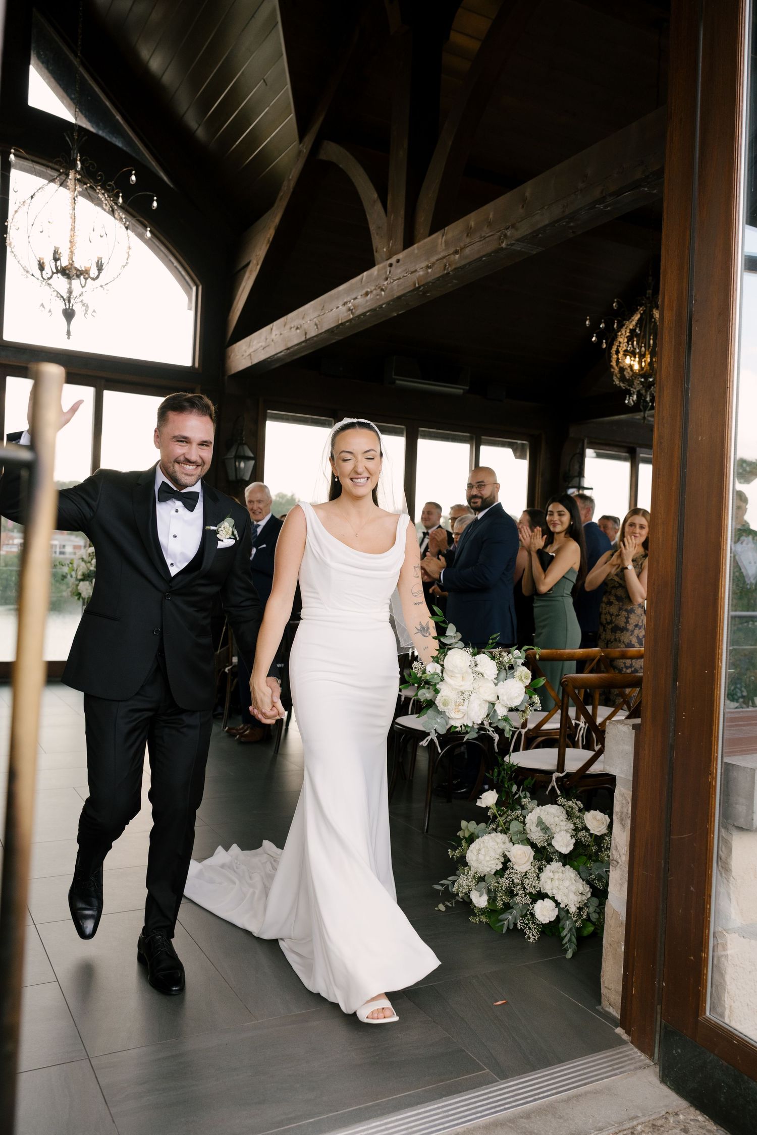 Newlyweds walking back down aisle hand-in-hand, bride in sleek white gown, groom in black tuxedo, guests applauding at Cambridge Mill venue.