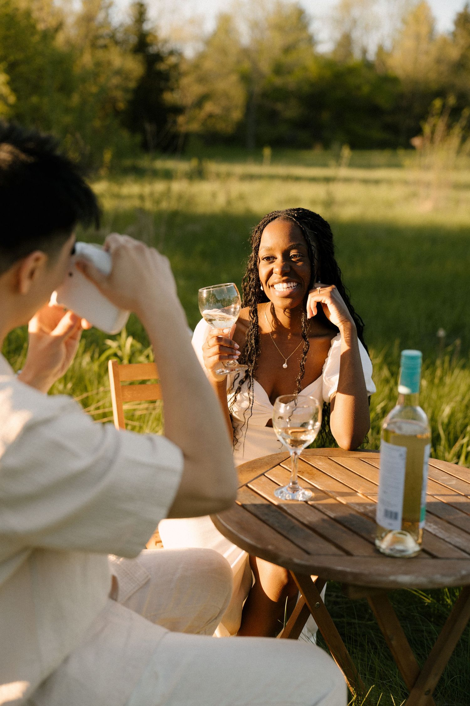 Man photographing smiling woman with wine glass at outdoor table in sunset.