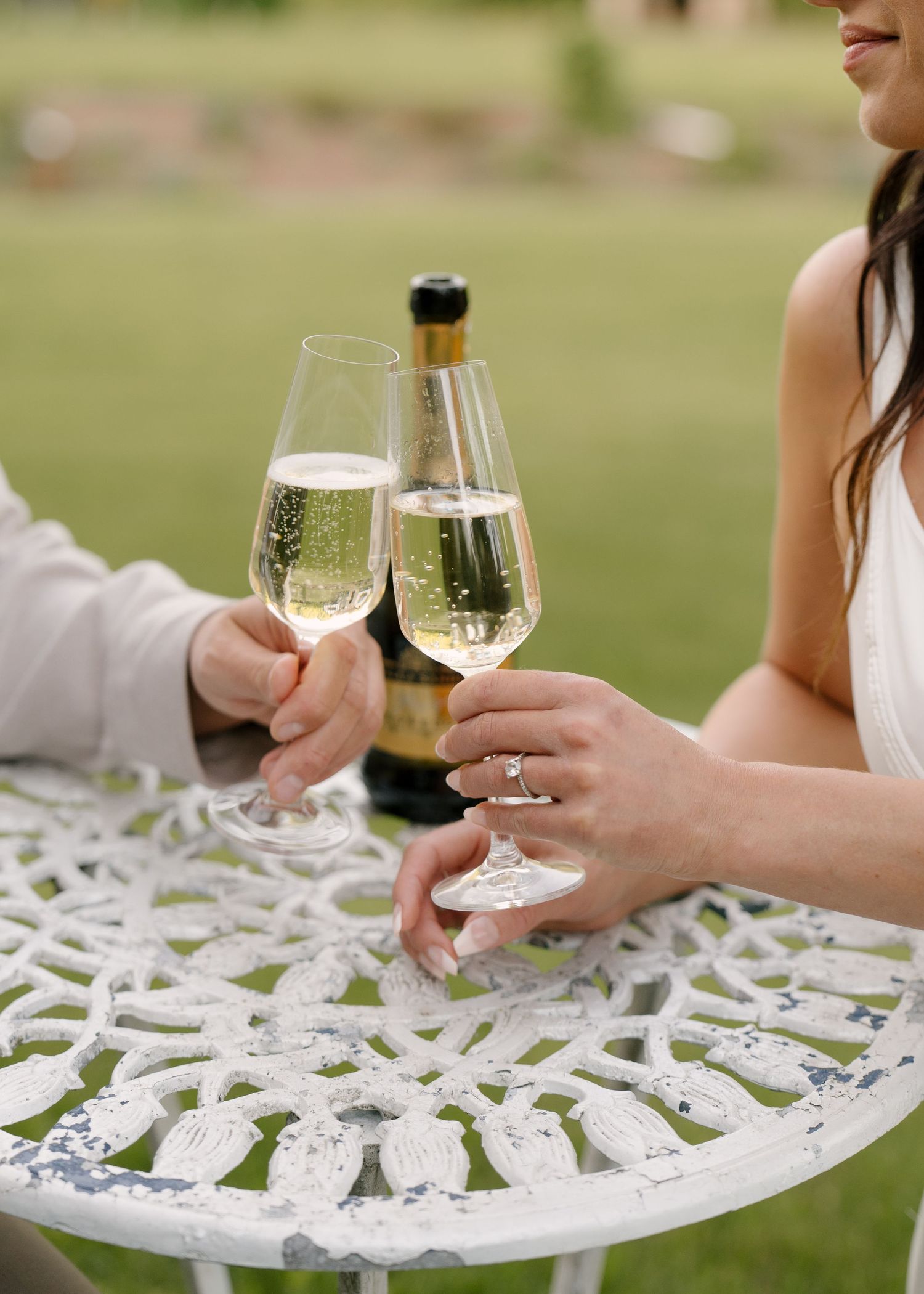 Couple toasting champagne glasses at ornate white iron table during outdoor wedding celebration.