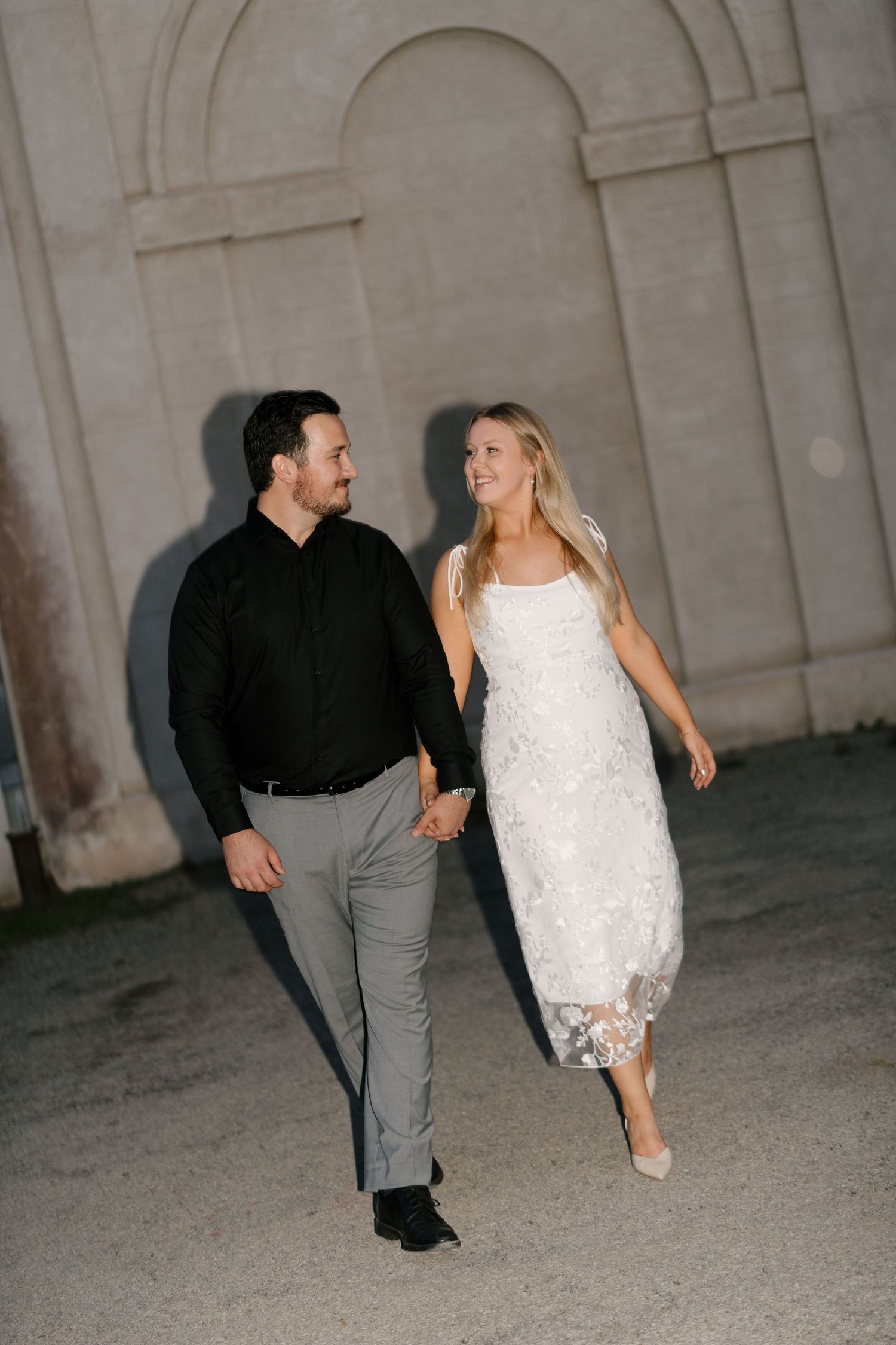 Smiling couple holding hands and walking at night near an illuminated stone architectural backdrop.